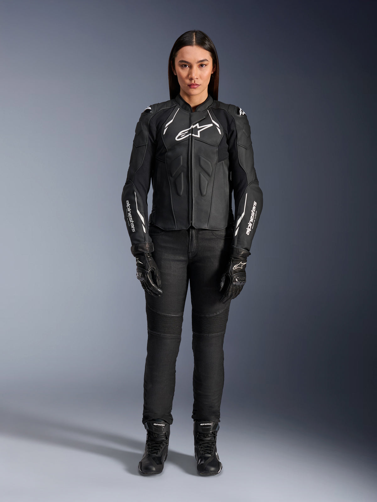 Giacca in pelle Alpinestars Stella Dusk da donna, nero e bianco, indossata da una modella con pantaloni e stivali neri coordinati, vista frontale, con loghi Alpinestars bianchi sul petto e sulle maniche, equipaggiamento protettivo in pregiata pelle bovina per donna
