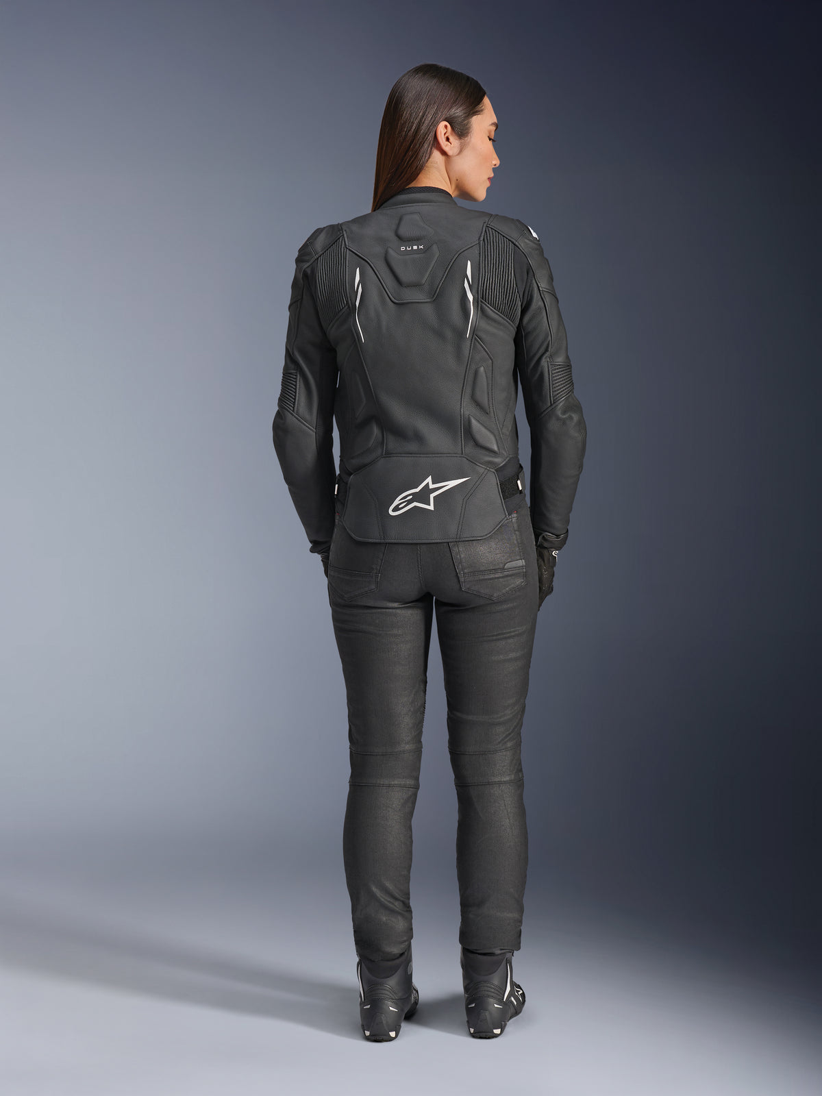 Giacca in pelle Alpinestars Stella Dusk da donna, nero, vista posteriore con logo Alpinestars bianco sulla parte bassa della schiena e marchio 'DUSK' sulla parte superiore, indossata con pantaloni e stivali da moto neri, mette in risalto la vestibilità femminile sagomata e la struttura protettiva in pelle