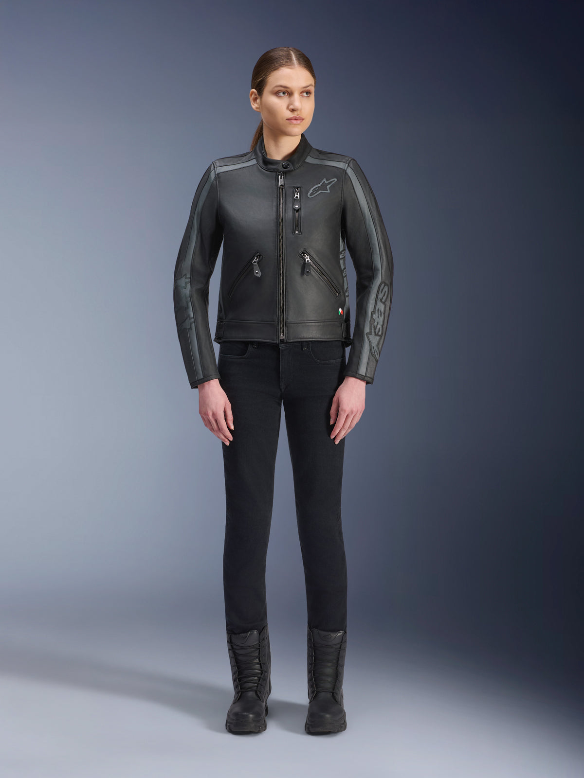 Alpinestars Stella Dyno Leather Jacket, giacca da moto da donna, nero, grigio catrame e grigio scuro, vista in studio a figura intera di una modella che indossa la giacca con pantaloni neri slim-fit e stivali neri, realizzata in pelle bovina con accenti grigi e logo Alpinestars sul petto e sulle maniche.
