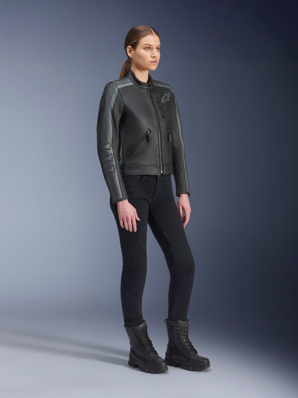 Alpinestars Stella Dyno Leather Jacket, giacca da moto da donna, nero Tar Gray e grigio scuro, indossata con pantaloni da moto e stivali neri, con strisce racing grigie sulle maniche e design in pelle heritage