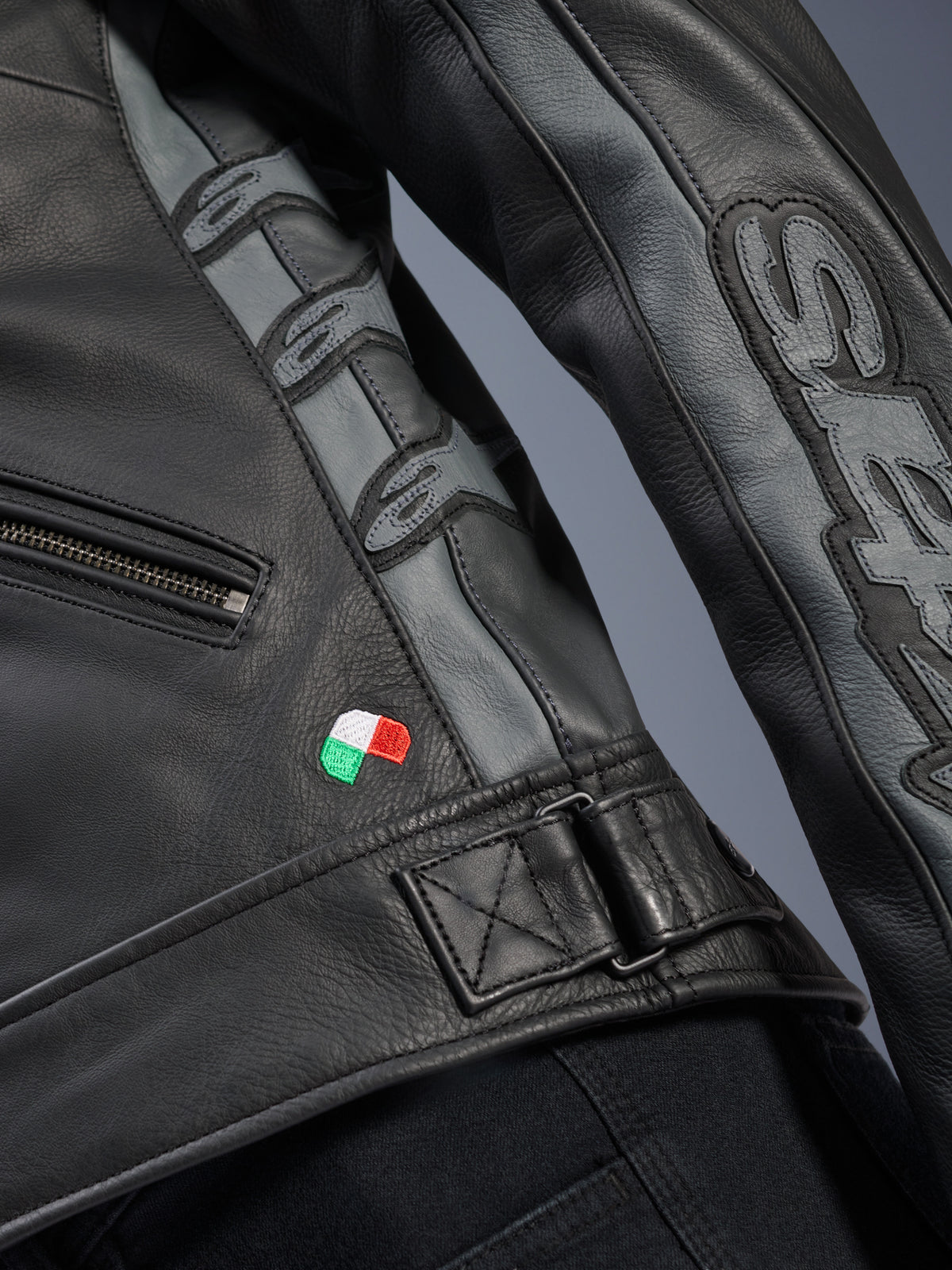 Dettaglio giacca in pelle Alpinestars Stella Dyno, pelle bovina nero e grigio fumo, primo piano con piccolo ricamo della bandiera italiana, fibbia di regolazione in vita e pannelli in pelle grigia con loghi Alpinestars in rilievo e marchio Stella sulla manica.
