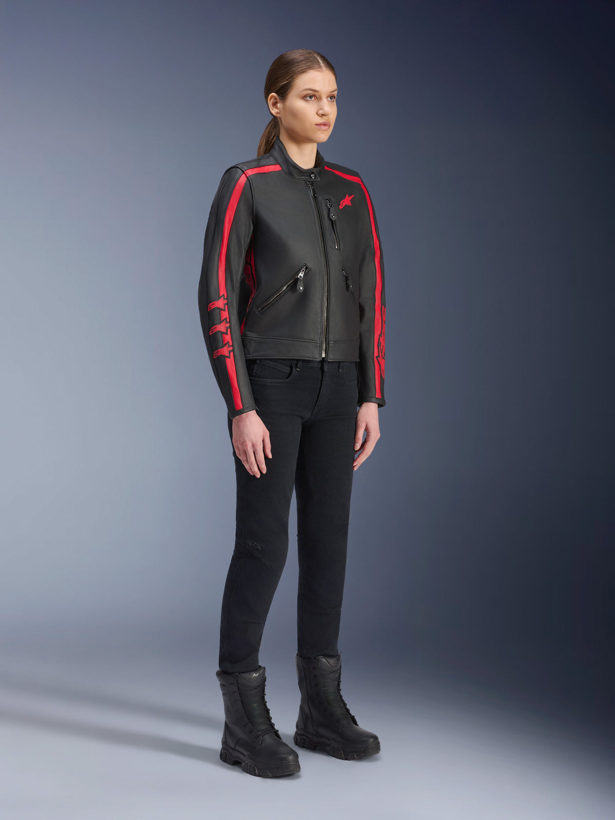 Modella che indossa la giacca in pelle Alpinestars Stella Dyno in Black Haute Red, abbinata a pantaloni e stivali neri, giacca da moto in pelle stile heritage con strisce rosse sulle braccia e logo rosso, abbigliamento da moto urbano e stradale per donna
