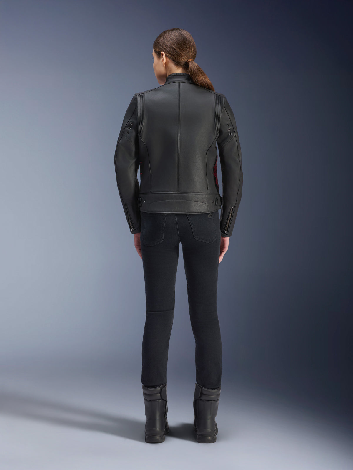 Vista posteriore di una donna che indossa la giacca in pelle Alpinestars Stella Dyno, nero con accenti Haute Red, abbinata a pantaloni neri slim-fit e stivali neri, design moderno heritage di giacca da moto in pelle.