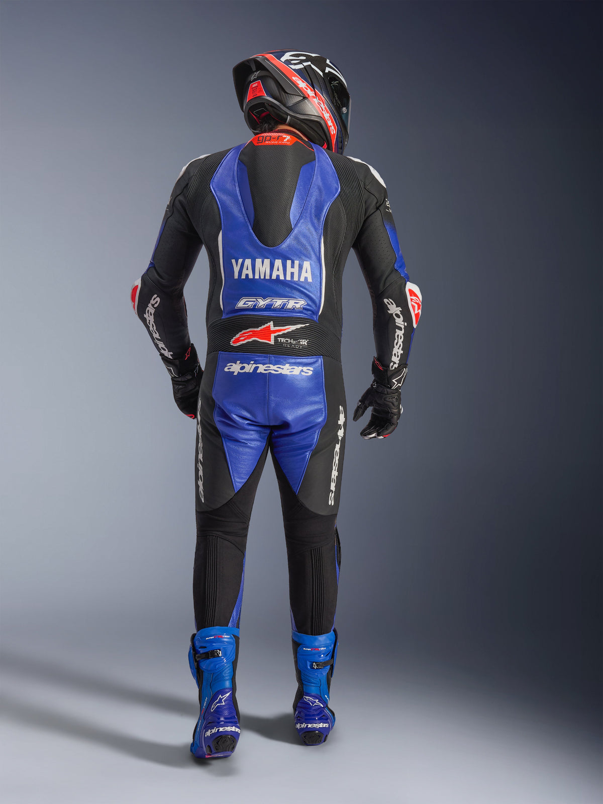 Tuta in pelle intera Alpinestars Yamaha GP-R7, vista posteriore di un pilota in equipaggiamento da gara completo, blu metallizzato e nero con accenti bianchi e rossi, loghi Yamaha e GYTR sulla gobba aerodinamica, indossata con stivali blu e casco da corsa