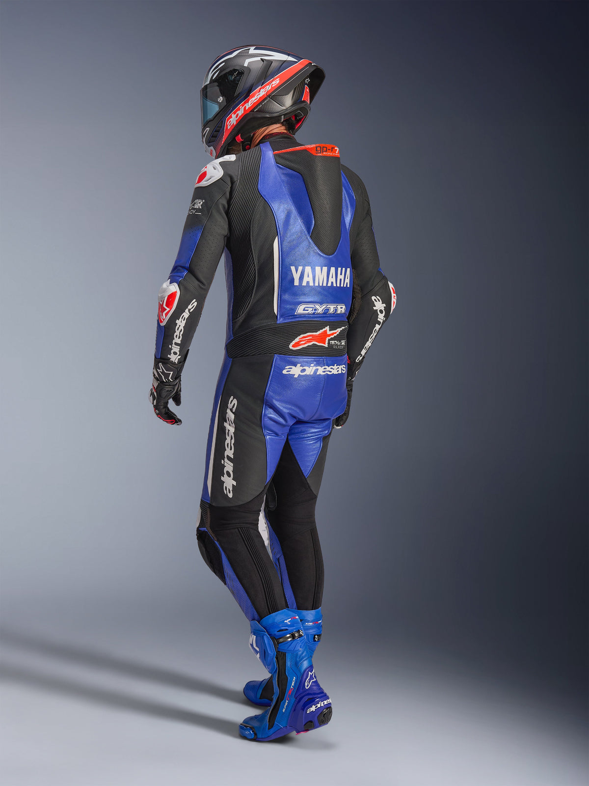 Tuta in pelle intera Alpinestars Yamaha GP-R7, blu metallizzato, nero e bianco, vista posteriore con gobba aerodinamica e marchio Yamaha, indossata con stivali e casco racing blu coordinati, tuta professionale in pelle per competizioni su pista