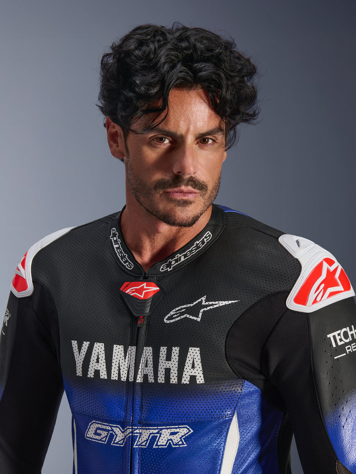 Ritratto in primo piano di un uomo che indossa la tuta in pelle intera Alpinestars Yamaha GP-R7, blu metallizzato e nero con loghi YAMAHA e GYTR bianchi, pelle traforata, slider sulle spalle bianchi con loghi a stella rossi, Tech-Air Ready, abbigliamento professionale per gare motociclistiche.