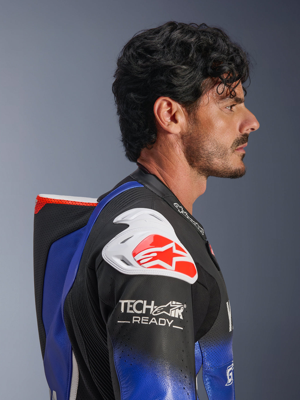 Tuta intera in pelle Alpinestars Yamaha GP-R7, blu metallizzato e nero con accenti bianchi e rossi, profilo laterale che mette in risalto la gobba aerodinamica e la protezione per la spalla bianca con logo Alpinestars rosso, marchio Tech-Air Ready sulla manica, pelle racing traforata di alta qualità.