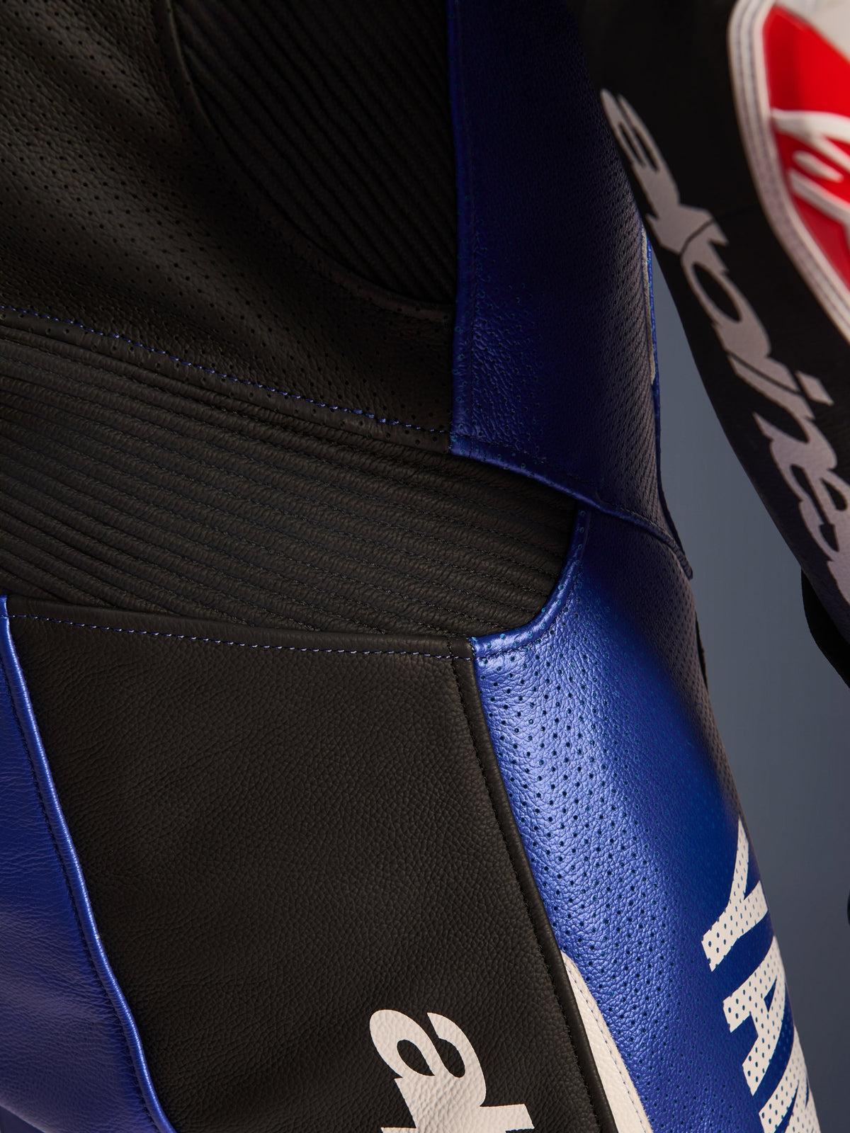 Dettaglio ravvicinato della tuta in pelle intera Alpinestars Yamaha GP-R7, blu metallizzato e nero, pannelli in pelle traforata per la ventilazione, inserti elastici a soffietto neri per la flessibilità e branding Yamaha bianco, costruzione in pelle bovina di qualità professionale.
