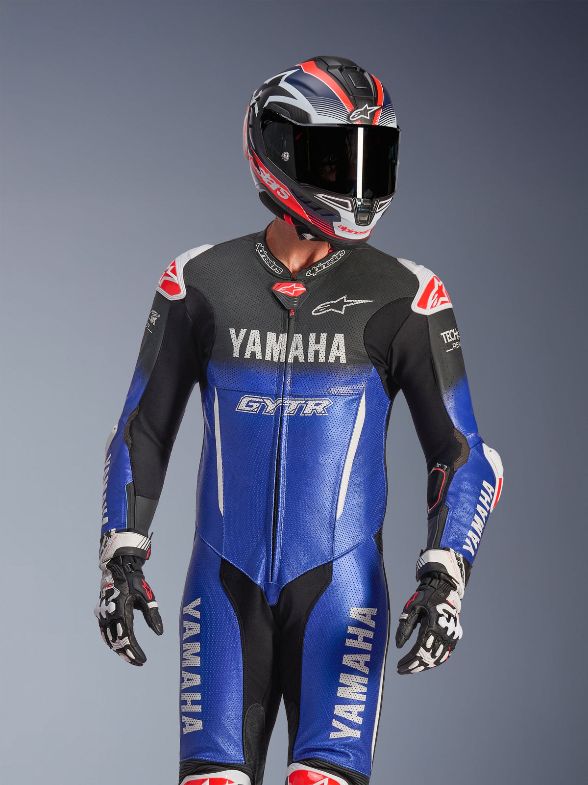 Tuta intera in pelle Yamaha GP-R7, blu metallizzato e nero con dettagli bianchi e rossi, indossata con casco e guanti da corsa coordinati, con marchi YAMAHA e GYTR, pannelli in pelle traforata e loghi Alpinestars, abbigliamento professionale da competizione motociclistica.