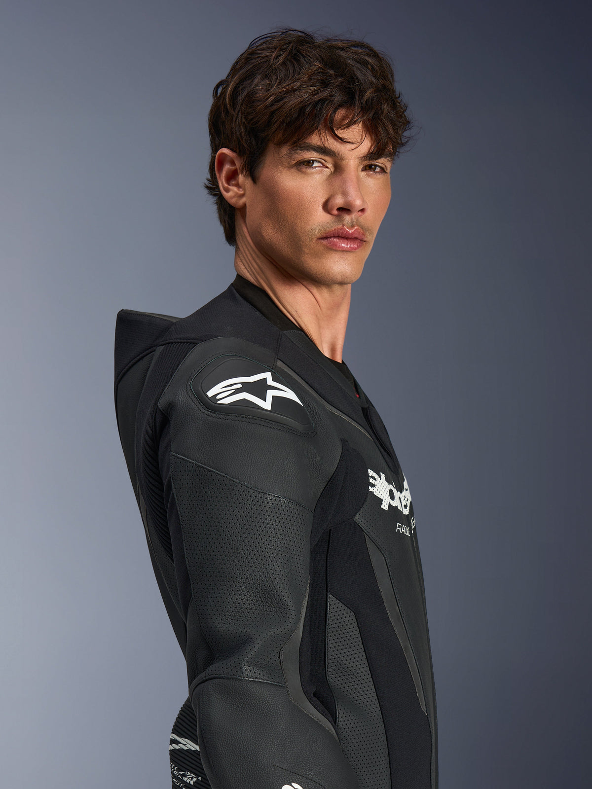 Modello uomo che indossa la tuta in pelle intera Alpinestars GP Force V2 in nero, vista di profilo che mette in risalto il logo a stella Alpinestars bianco sulla protezione per le spalle DFS Lite, i pannelli in pelle traforata e la gobba aerodinamica.