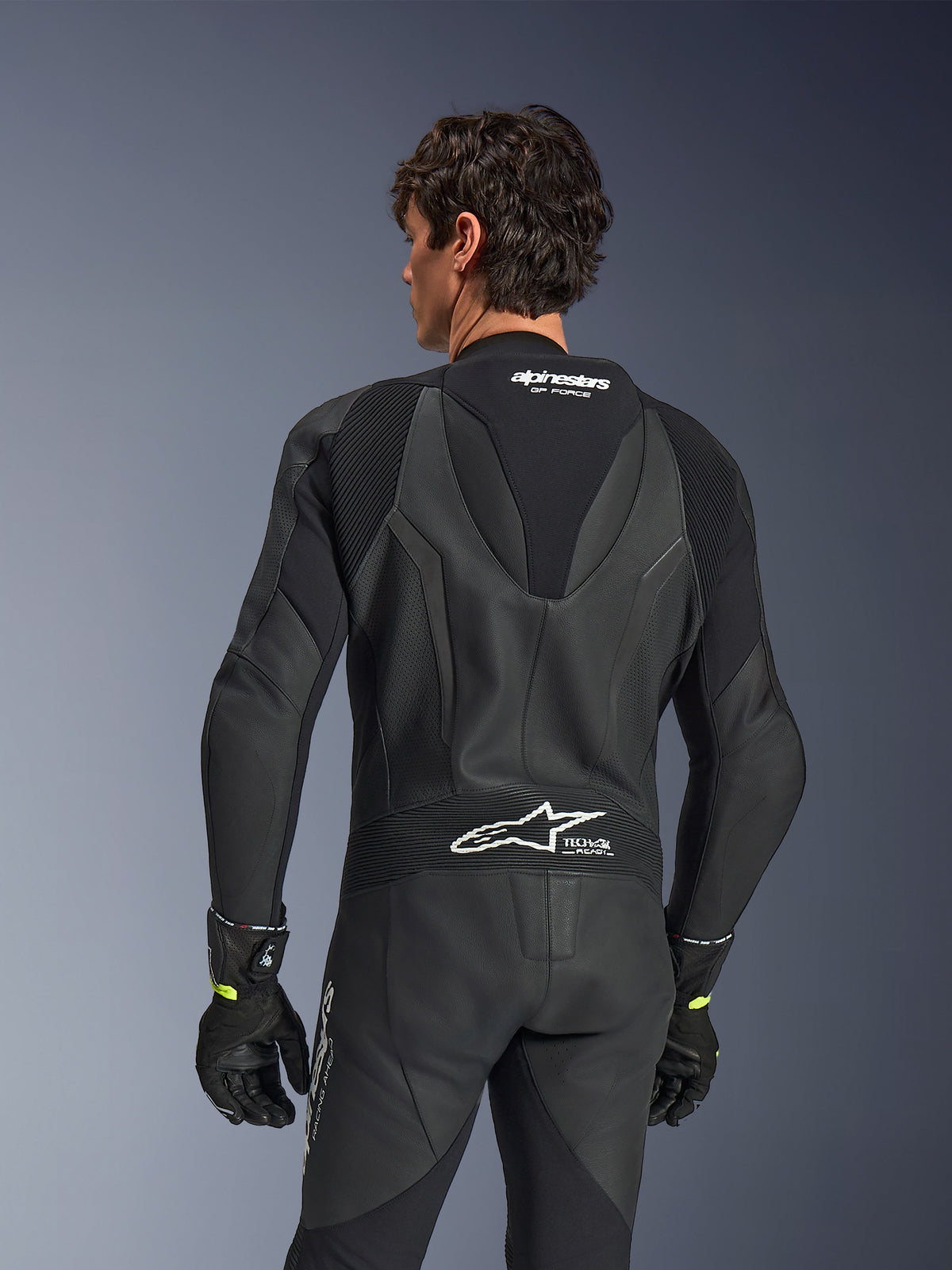 Tuta in pelle Alpinestars GP Force V2, intera, nera, vista posteriore con pelle traforata e pannelli elasticizzati a fisarmonica, loghi bianchi Alpinestars e Tech-Air Ready sulla parte superiore e inferiore della schiena, indossata con guanti neri, abbigliamento da competizione motociclistica ad alte prestazioni