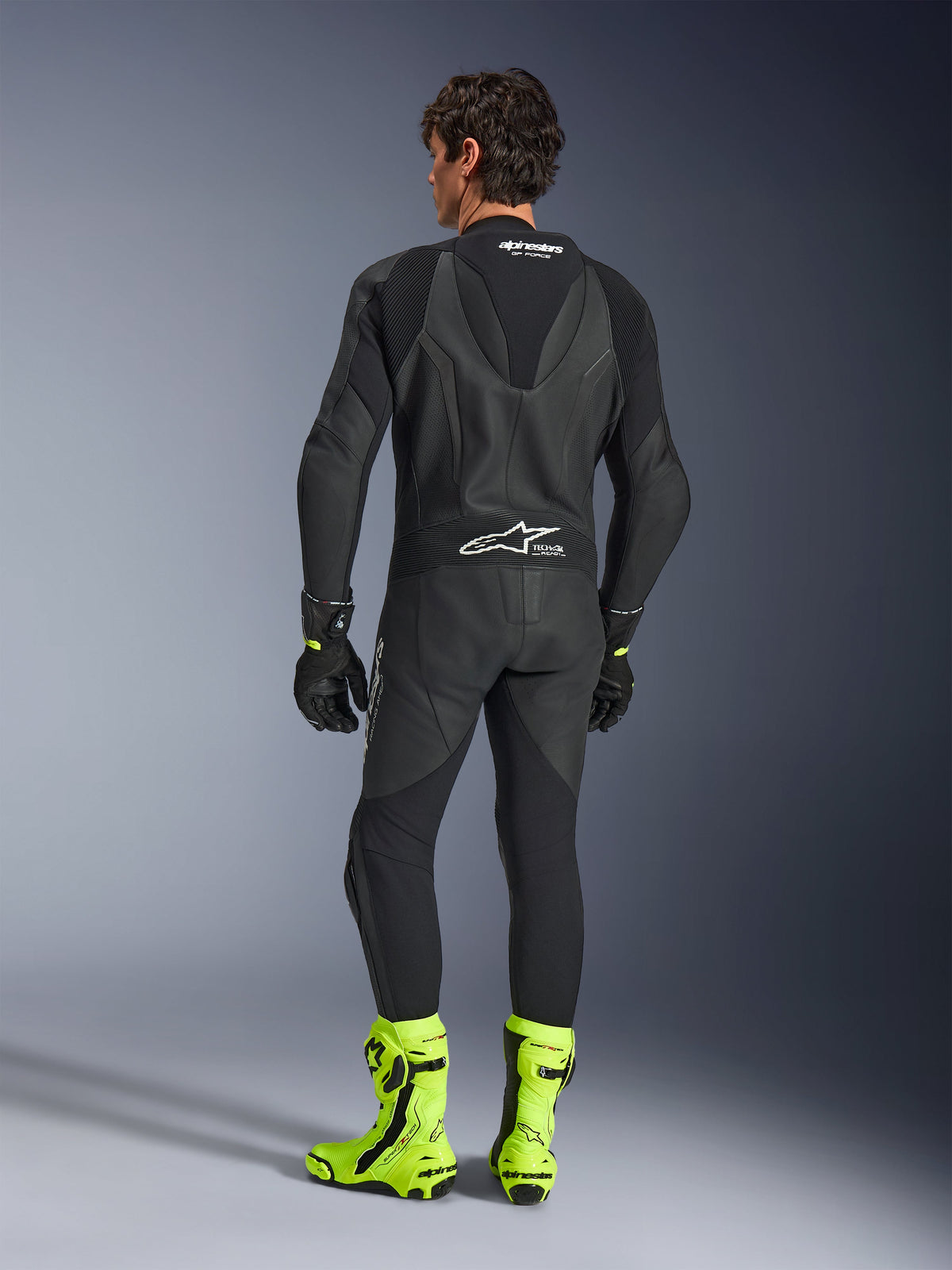 Alpinestars GP Force V2 Leather Suit - 1 pezzo, nero, vista posteriore indossata da un modello, con gobba aerodinamica e marchio Tech-Air Ready, abbinata a guanti neri e stivali da corsa giallo fluorescente, equipaggiamento professionale da pista.