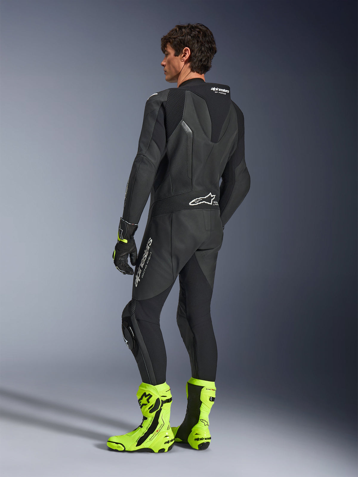 Tuta in pelle Alpinestars GP Force V2, tuta da moto racing monopezzo nera, vista posteriore che mostra la gobba aerodinamica e la pelle traforata, indossata con stivali da corsa giallo neon e guanti neri, attrezzatura professionale pronta per la pista