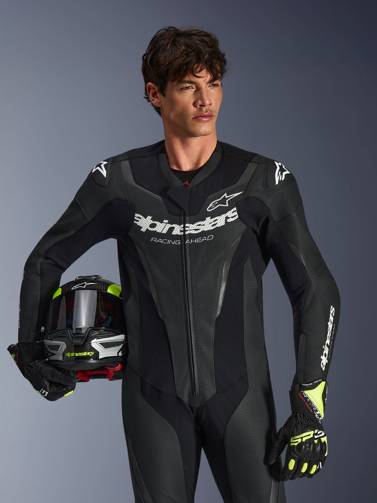 Tuta in pelle Alpinestars GP Force V2, tuta da moto intera nera con pannelli in pelle traforata e loghi bianchi, indossata da un modello che tiene un casco e guanti abbinati, illuminazione da studio