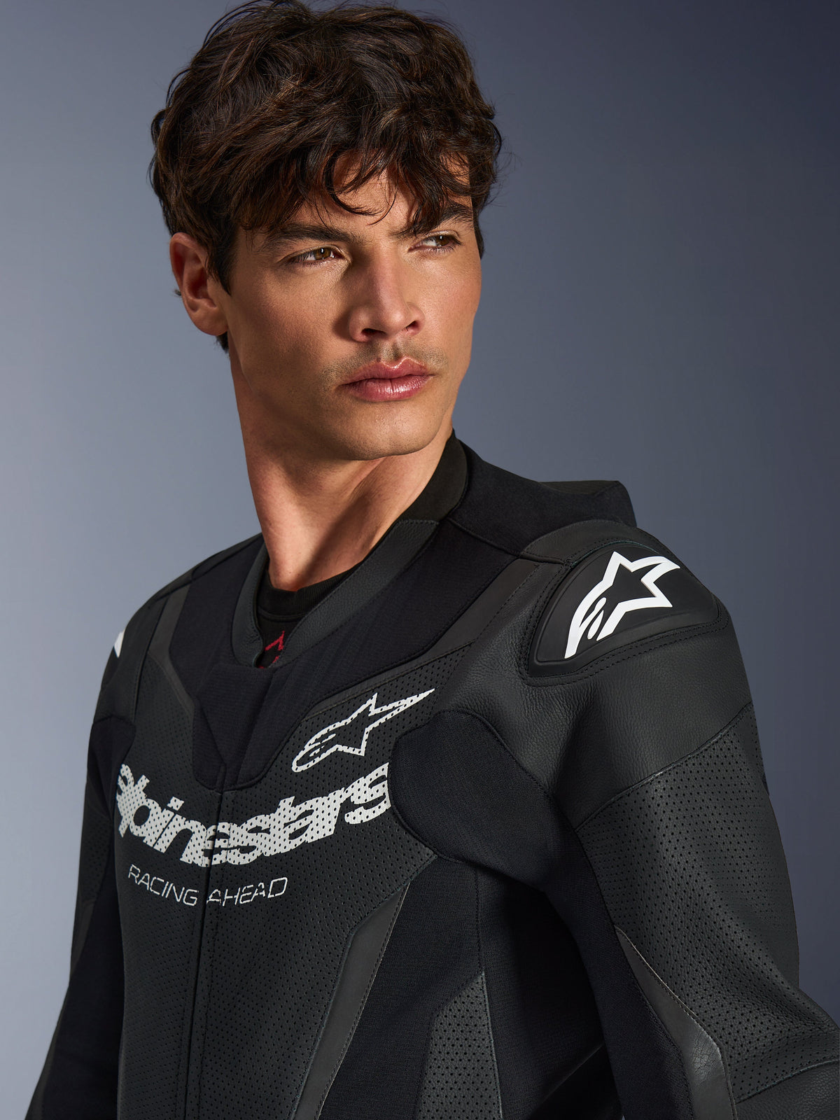 Primo piano di un uomo che indossa la tuta in pelle Alpinestars GP Force V2 in nero, caratterizzata da una struttura in pelle traforata, logo Alpinestars con scritta 'RACING AHEAD' sul petto e logo a stella bianco sugli slider delle spalle.