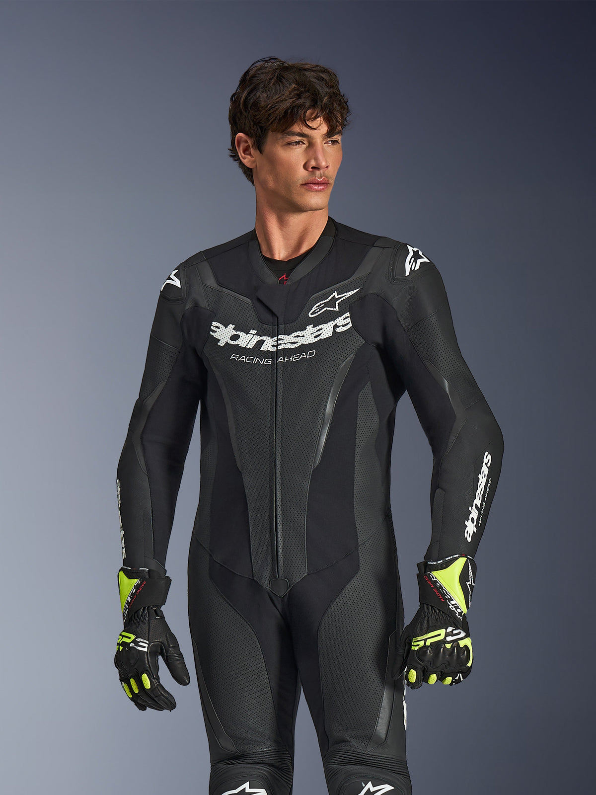 Tuta in pelle Alpinestars GP Force V2 1-Piece, nero, branding e logo Alpinestars bianchi, pannelli in pelle traforata per la ventilazione, indossata da un modello con guanti neri e giallo neon, abbigliamento da moto ad alte prestazioni