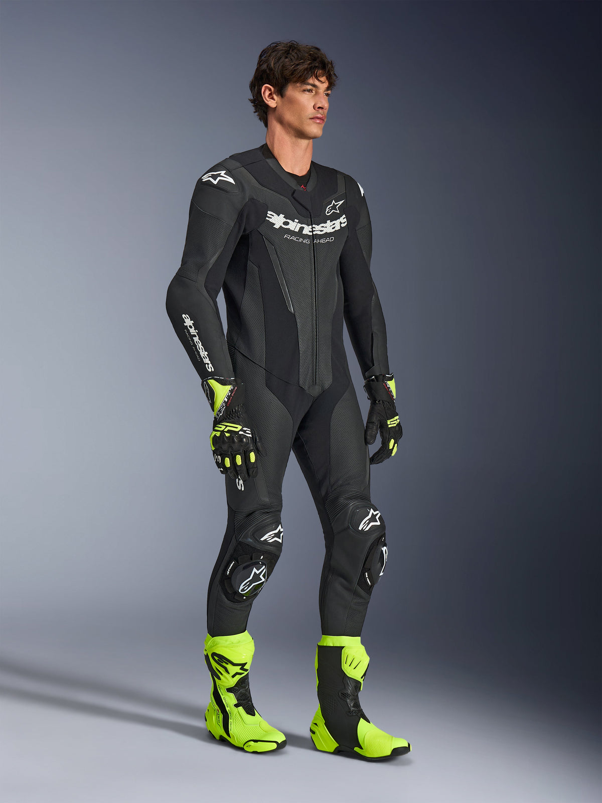 Modello che indossa la tuta in pelle intera Alpinestars GP Force V2 nera, con loghi Alpinestars bianchi, sezioni in pelle traforata e protezioni integrate, abbinata a stivali e guanti da gara giallo fluo e neri