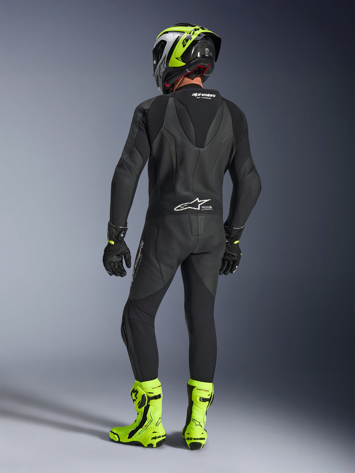 Vista posteriore di un pilota che indossa la tuta in pelle monopezzo Alpinestars GP Force V2 in nero, che mostra la gobba aerodinamica e il marchio Tech-Air Ready, abbinata a un casco nero e giallo fluorescente e stivali da corsa giallo acceso.