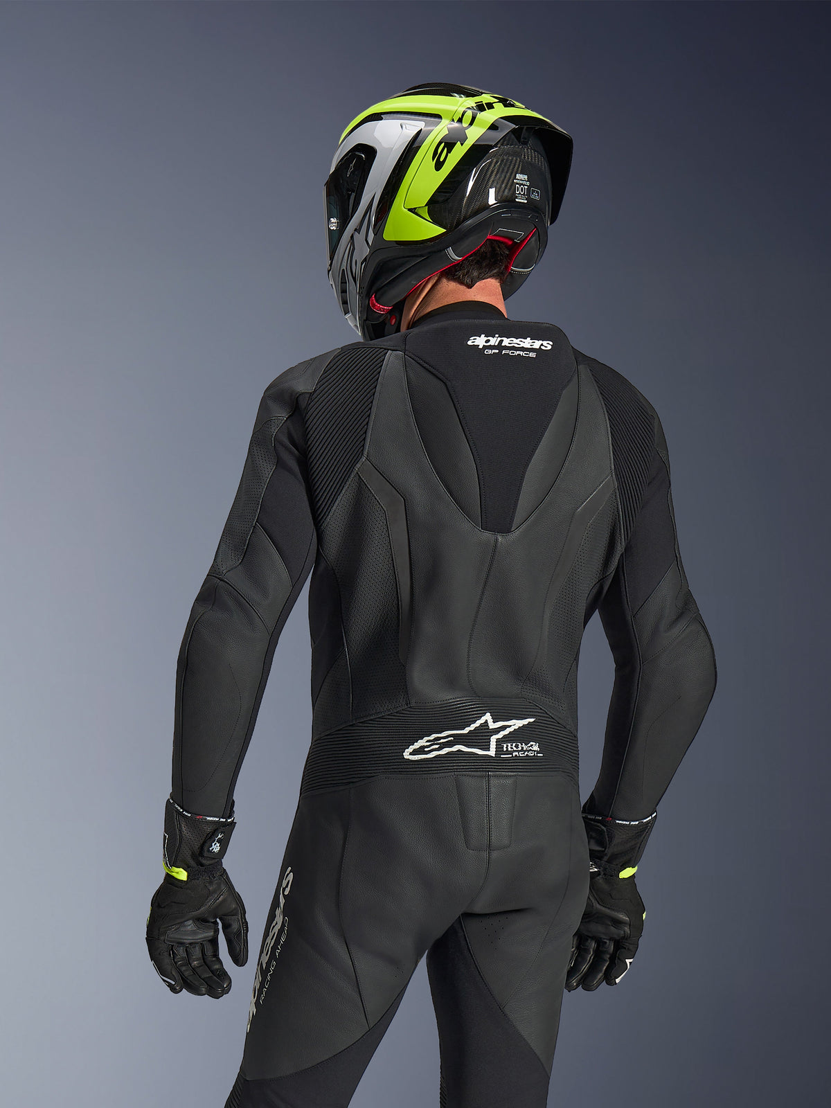 Alpinestars GP Force V2 Leather Suit 1-Piece, nero, vista posteriore che mostra la gobba aerodinamica e la struttura in pelle traforata, indossata con casco da corsa nero e giallo neon e guanti neri, dotata di pannelli elasticizzati a fisarmonica e logo Tech-Air Ready bianco.