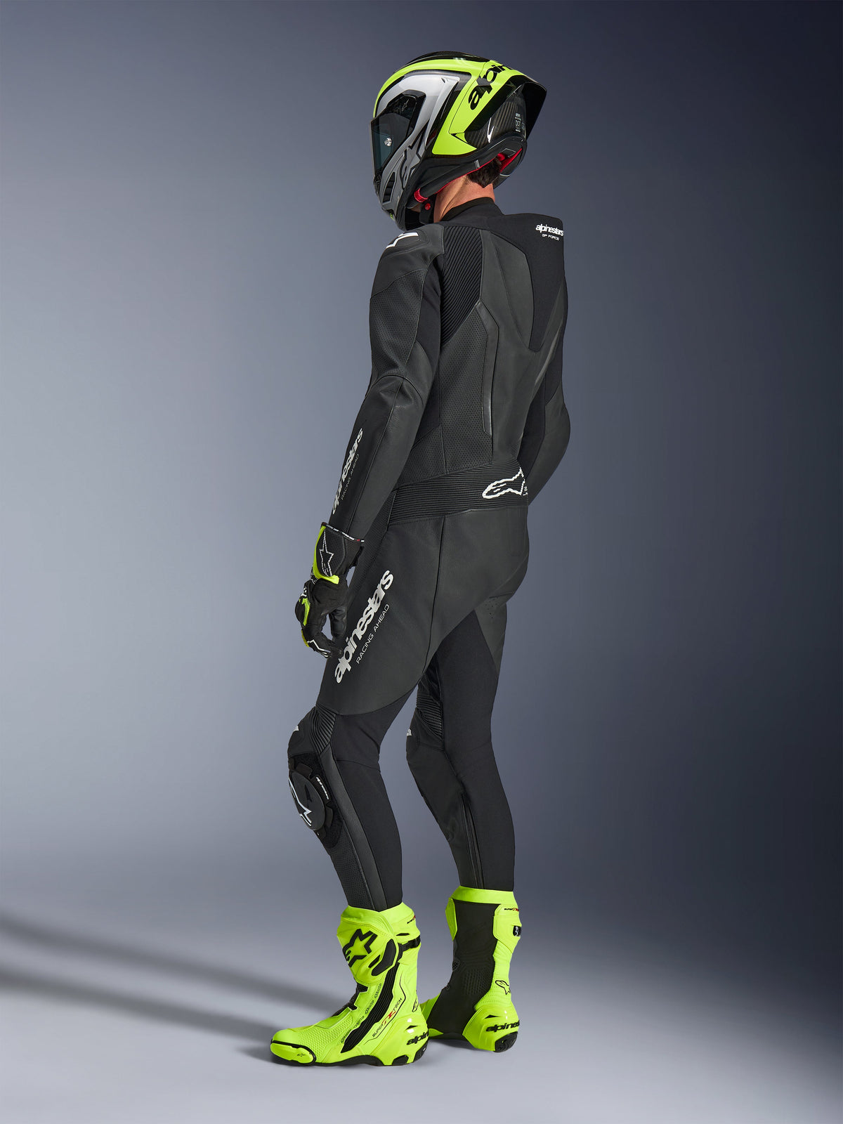 Pilota di moto con tuta in pelle Alpinestars GP Force V2 1-Piece in nero, vista posteriore laterale con loghi bianchi su coscia e parte bassa della schiena, gobba aerodinamica, abbinata a casco nero e giallo neon, guanti e stivali racing coordinati, abbigliamento professionale da pista e guida sportiva.
