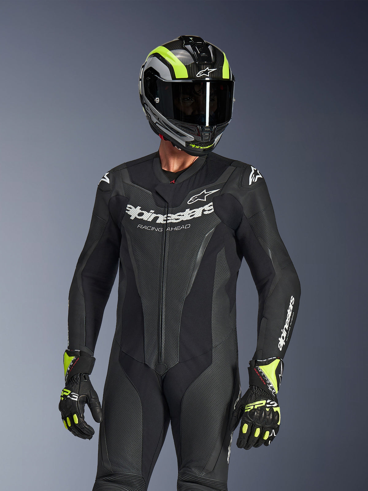 Tuta in pelle monopezzo Alpinestars GP Force V2, nera, indossata da un pilota con casco e guanti neri e giallo neon, logo Alpinestars bianco sul petto, pannelli in pelle traforata per la ventilazione e design professionale da gara.