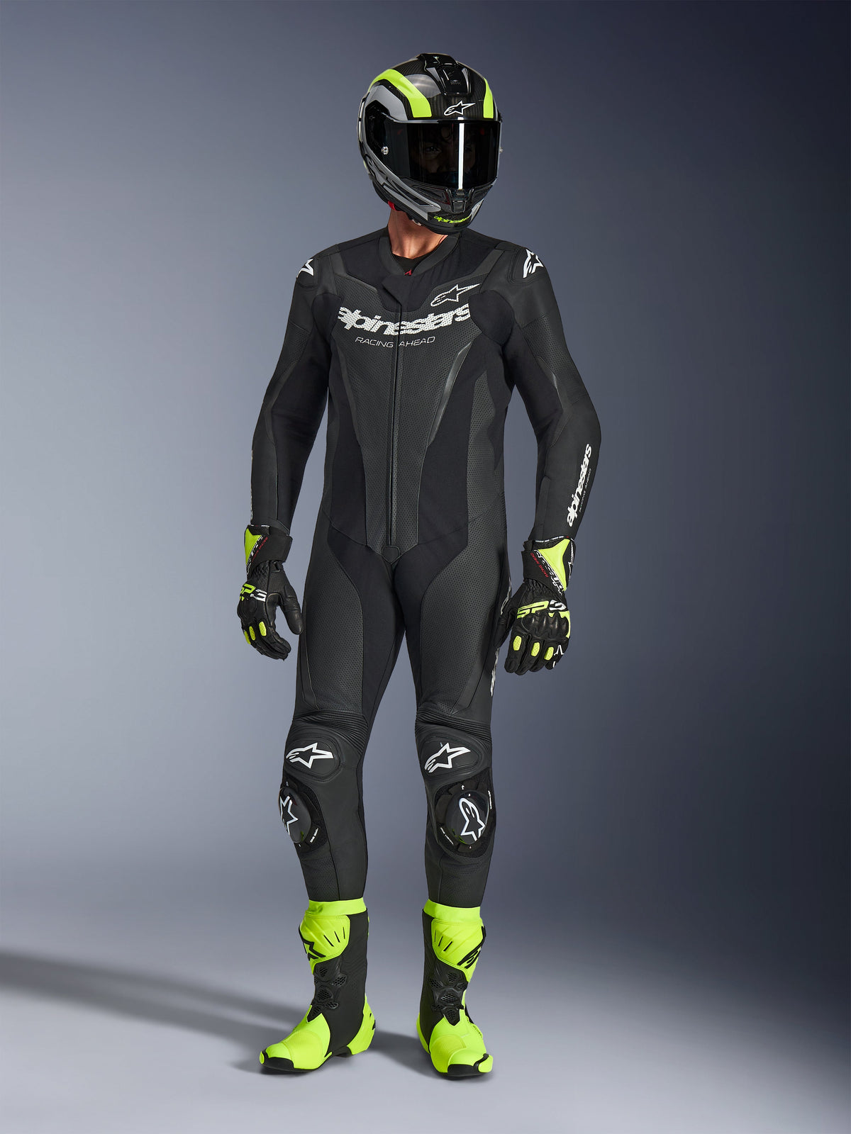 Pilota di moto che indossa la tuta in pelle Alpinestars GP Force V2 in nero, tuta intera con loghi bianchi, abbinata a casco racing, guanti e stivali nero e giallo fluo, abbigliamento professionale da pista su sfondo grigio da studio
