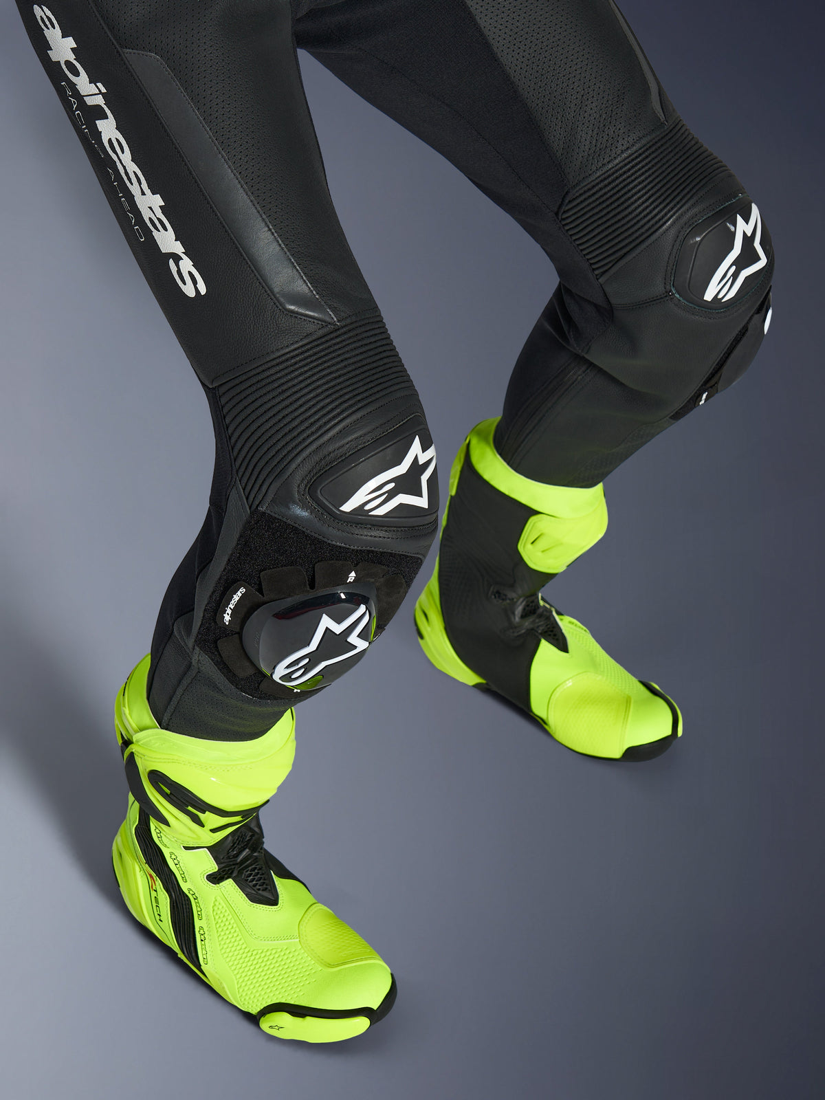 Tuta in pelle Alpinestars GP Force V2, tuta in pelle nera monopezzo, primo piano di gambe e ginocchia con saponette con logo bianco, pannelli elasticizzati a fisarmonica, indossata con stivali da corsa giallo neon brillante, equipaggiamento da corsa motociclistica ad alte prestazioni