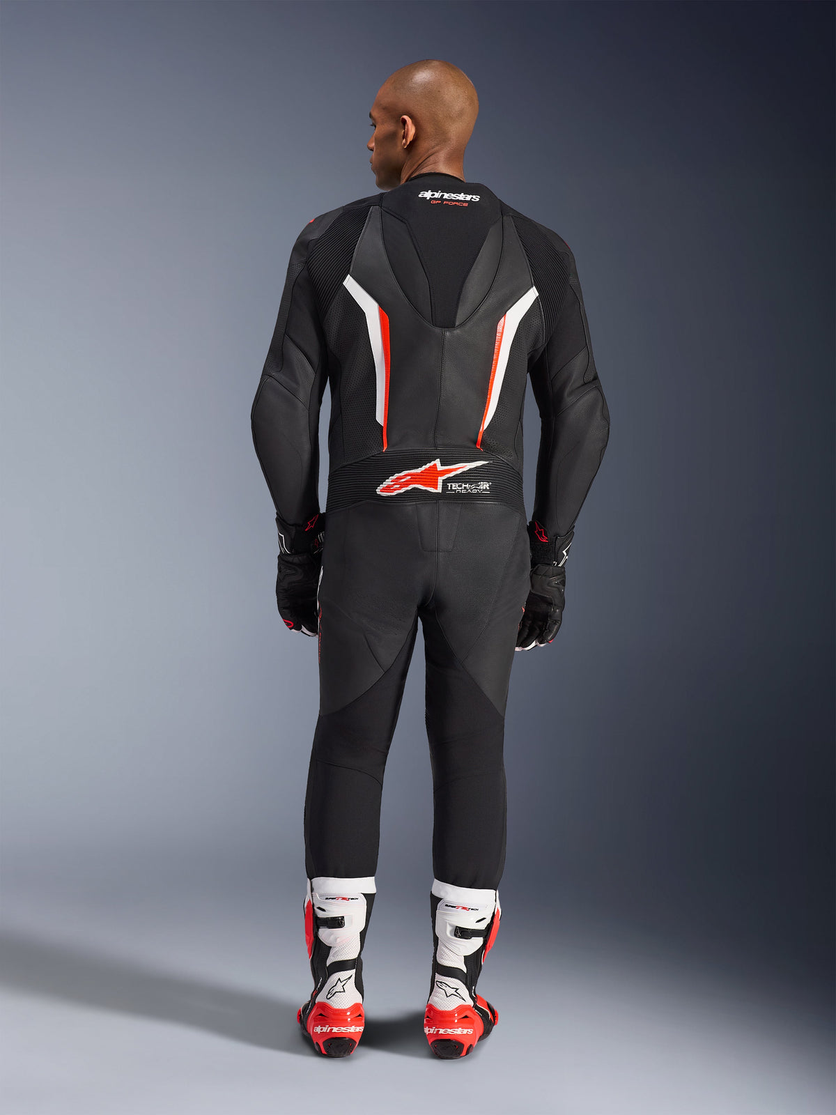 Vista posteriore di un modello che indossa la tuta in pelle intera Alpinestars GP Force V2 in Nero Bianco Rosso Fluo, mettendo in evidenza la gobba aerodinamica e il marchio Tech-Air Ready, abbinata a stivali da corsa professionali bianchi e rossi e guanti neri.