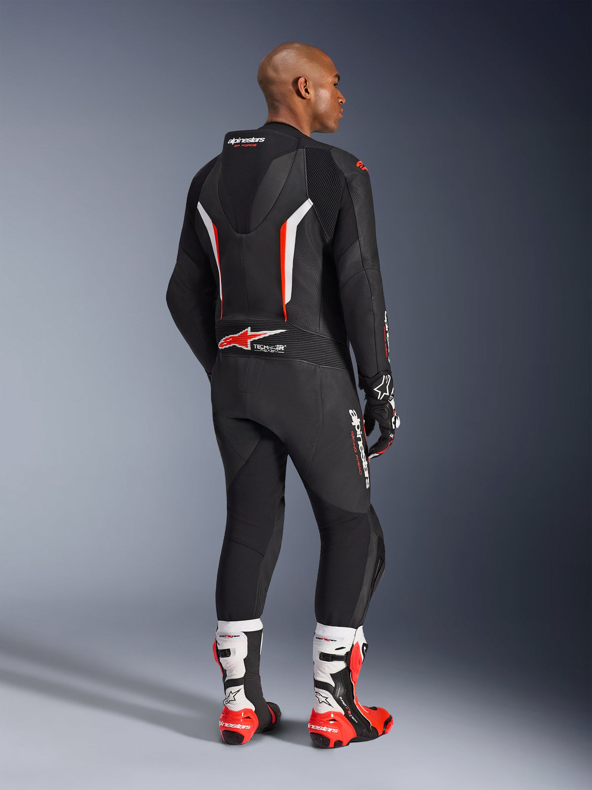 Pilota che indossa la tuta in pelle intera GP Force V2, nero con accenti bianco e rosso fluo, visto dal retro che mostra la gobba aerodinamica e il marchio Alpinestars, abbinato a guanti da corsa coordinati e stivali bianchi e rossi, equipaggiamento professionale da moto