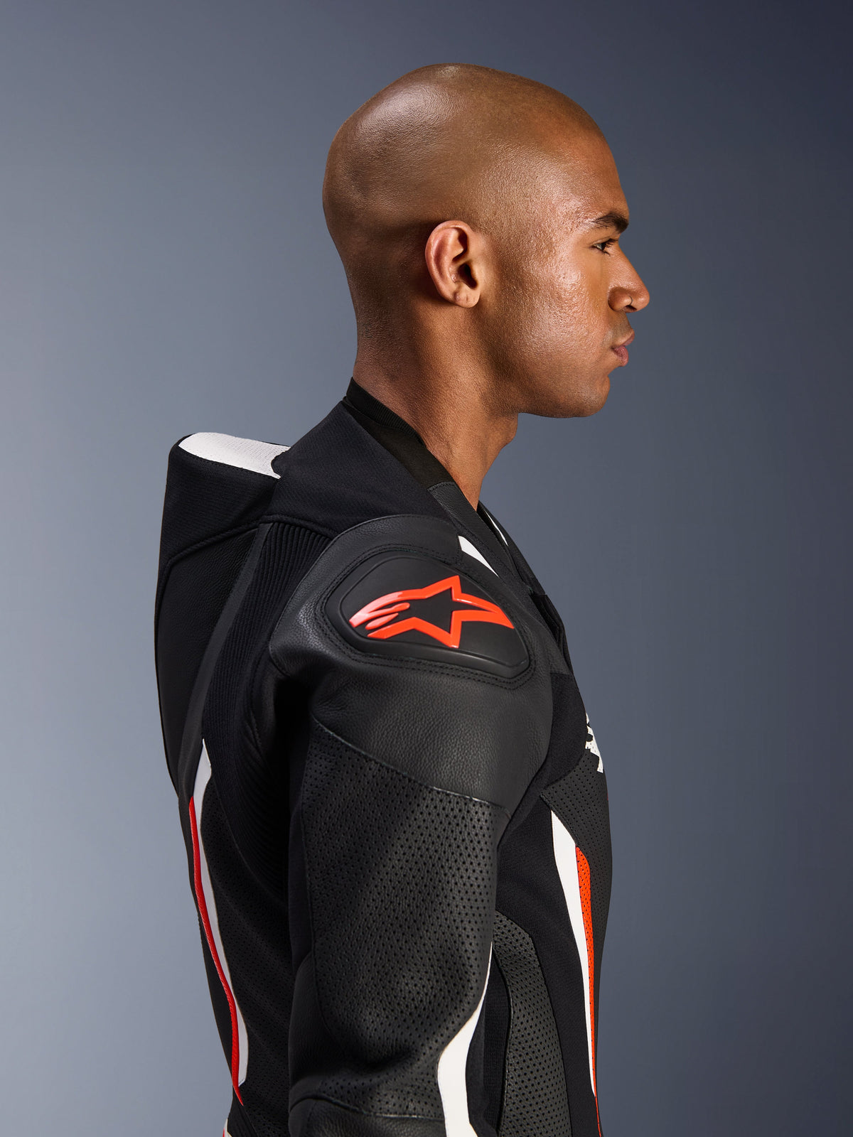 Alpinestars GP Force V2 Leather Suit - 1Piece, profilo laterale di un pilota, colorazione Nero Bianco Rosso Fluo, che mette in risalto la gobba aerodinamica, le zone in pelle traforata e la protezione per le spalle DFS Lite con logo Alpinestars rosso