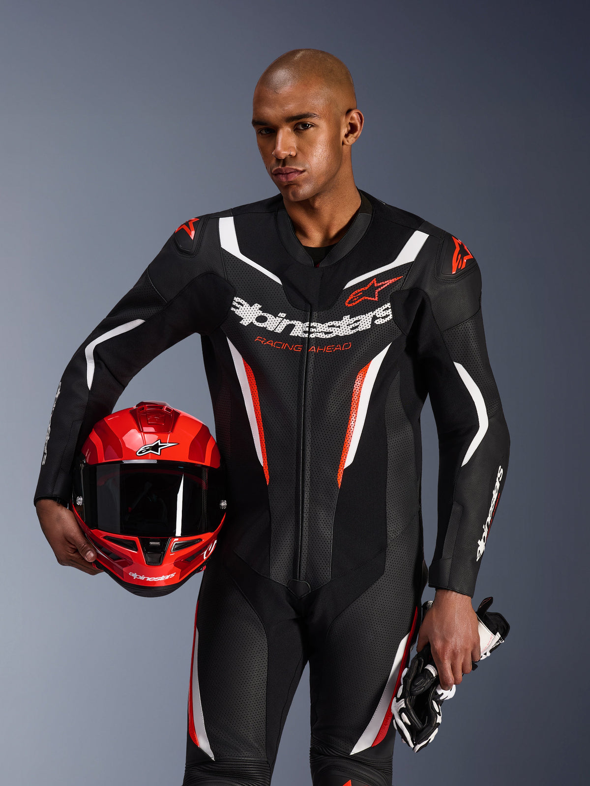 Alpinestars GP Force V2 Leather Suit, tuta da moto intera, nero, bianco e rosso fluo, indossata da un modello che tiene un casco rosso e guanti, design racing con pelle traforata e loghi evidenti