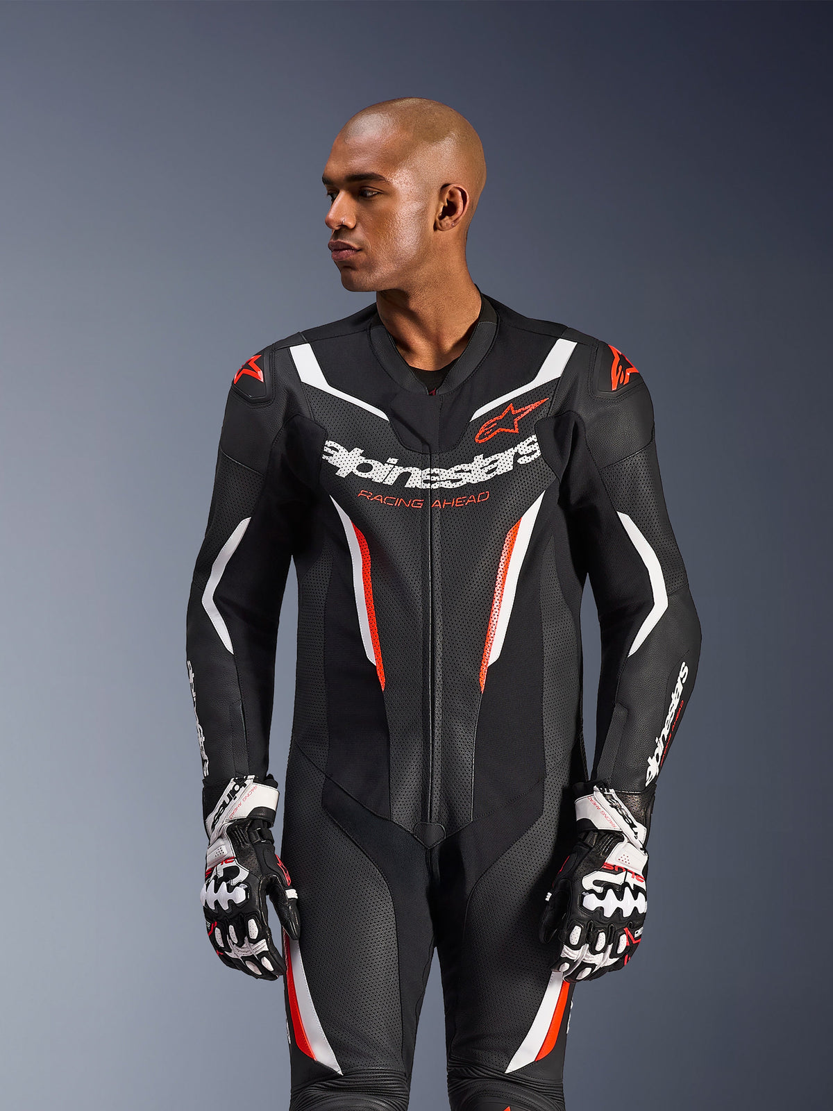 Tuta in pelle Alpinestars GP Force V2 1-Piece, nero bianco rosso fluo, indossata da un modello con guanti da corsa, con struttura in pelle traforata, vivaci accenti grafici bianchi e rossi e marchio Alpinestars sul petto.