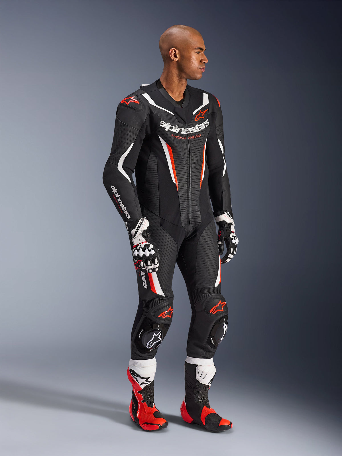 Modello che indossa la tuta in pelle intera Alpinestars GP Force V2 in Nero Bianco Rosso Fluo, abbigliamento da corsa ad alte prestazioni con pannelli in pelle traforata e scudi DFS Lite, abbinata a stivali e guanti racing coordinati rossi e bianchi, estetica motorsport professionale.