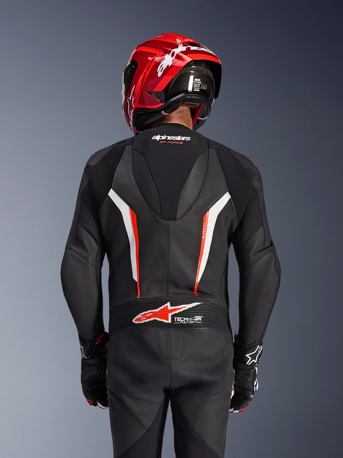 Tuta in pelle intera Alpinestars GP Force V2, nero, bianco e rosso fluo, vista posteriore del pilota con casco da corsa rosso e guanti, con pannelli in pelle traforata, gobba aerodinamica con marchio Alpinestars e logo Tech-Air Ready sulla parte bassa della schiena, abbigliamento professionale da gara motociclistica.