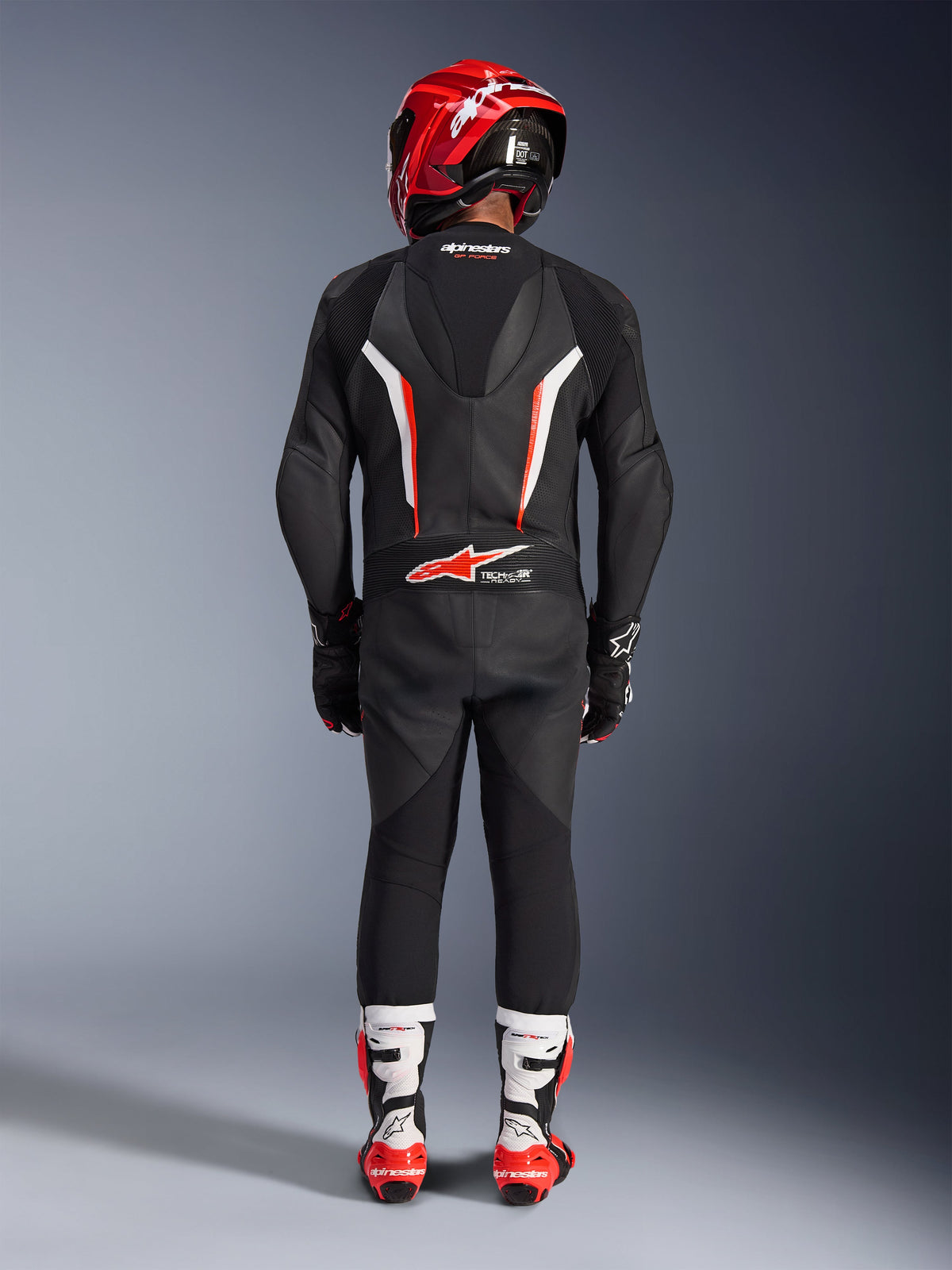 Pilota visto da dietro con tuta in pelle Alpinestars GP Force V2 1-Piece in nero, bianco e rosso fluo, caratterizzata da gobba aerodinamica e marchio Tech-Air Ready, coordinata con casco racing rosso e stivali bianchi e rossi, design professionale per attrezzatura da pista.