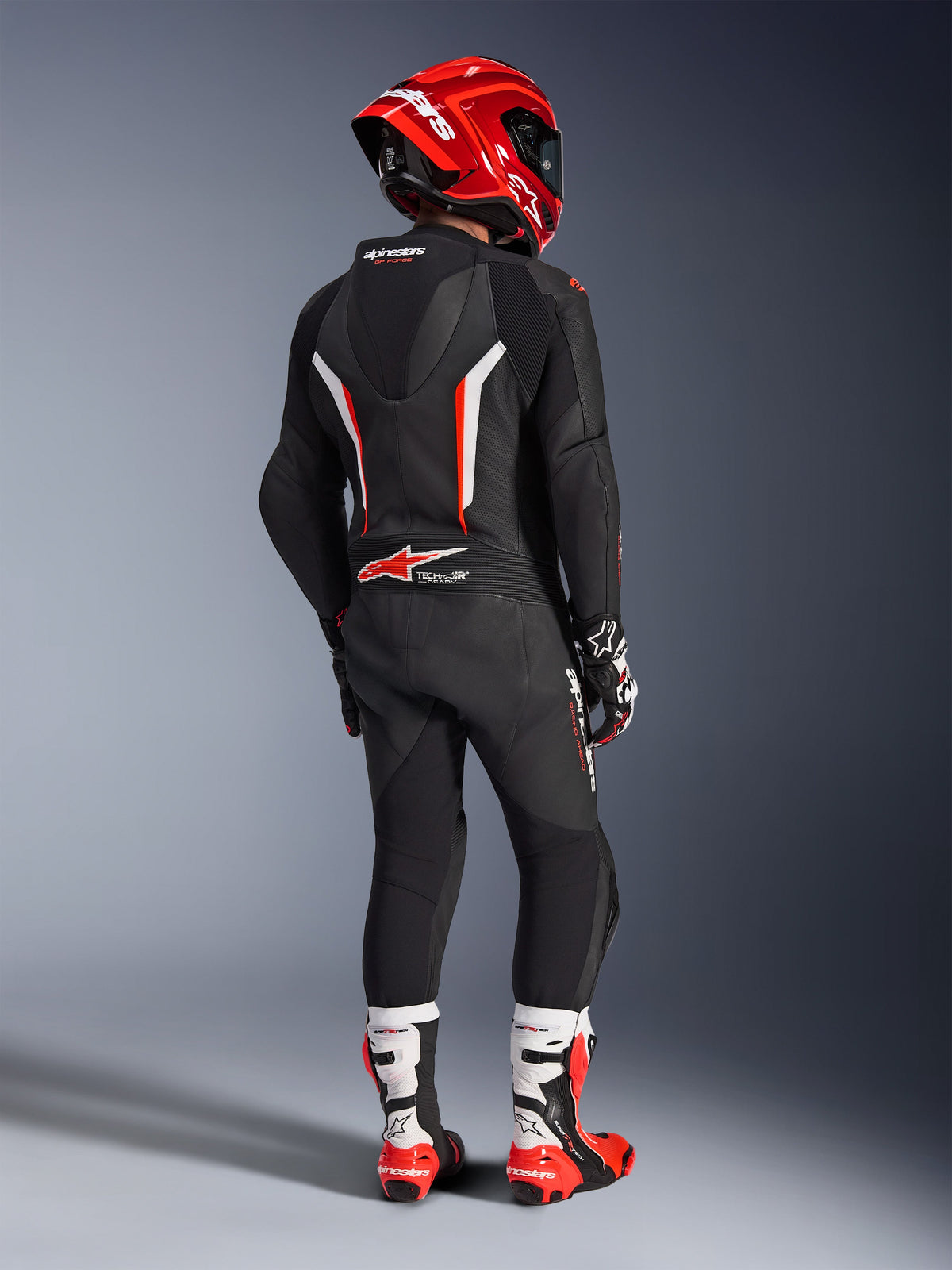 Pilota visto da dietro che indossa la tuta in pelle intera Alpinestars GP Force V2 in nero, bianco e rosso fluo, con gobba aerodinamica e pannelli in pelle traforata, abbinata a un casco Alpinestars rosso, guanti racing e stivali da moto rossi e bianchi, abbigliamento professionale da gara su pista.