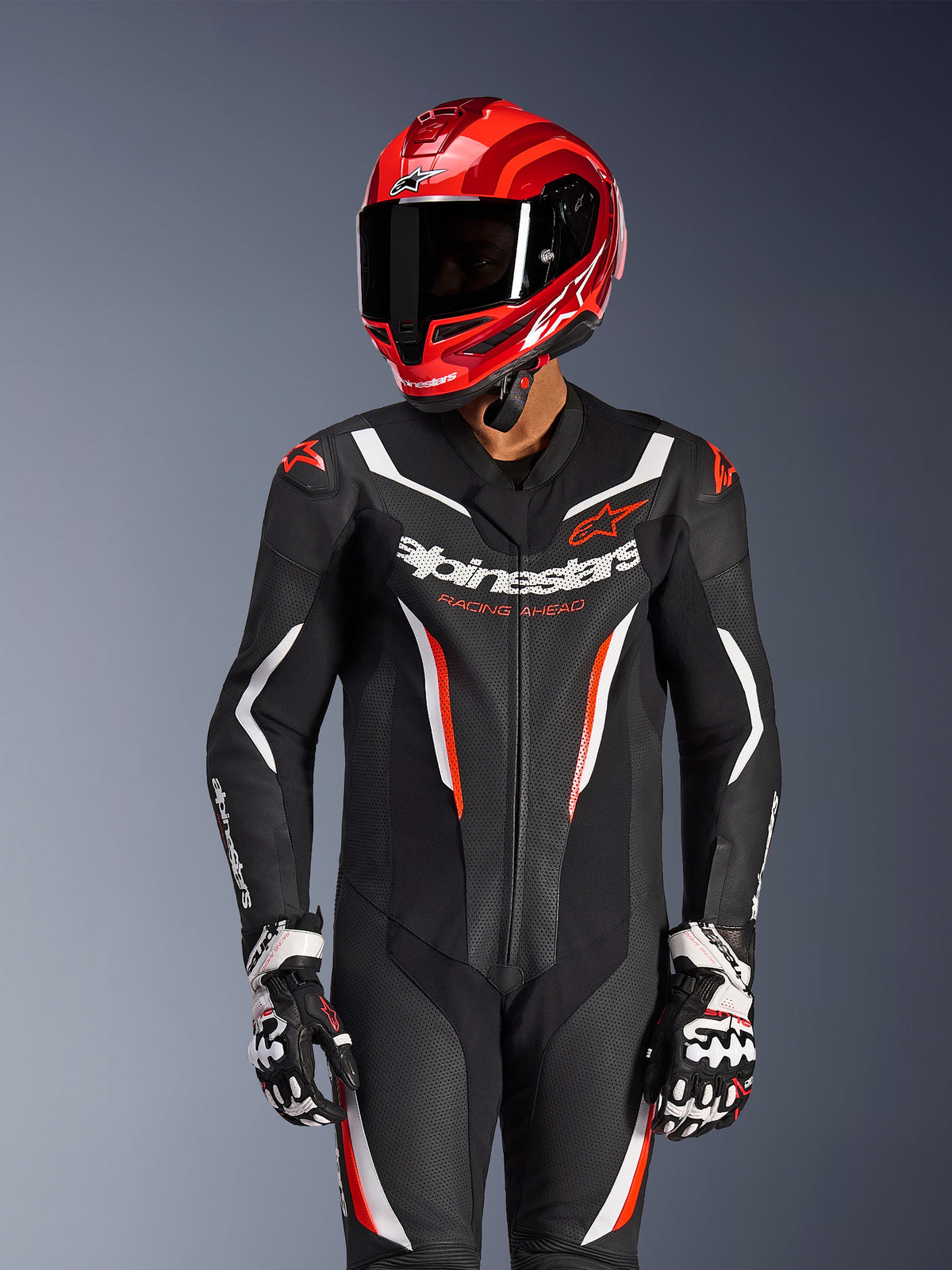 Pilota di moto che indossa la tuta in pelle intera Alpinestars GP Force V2 in nero, bianco e rosso fluo, casco da corsa e guanti rossi coordinati, dotata di pannelli in pelle traforata e branding audace, equipaggiamento racing ad alte prestazioni