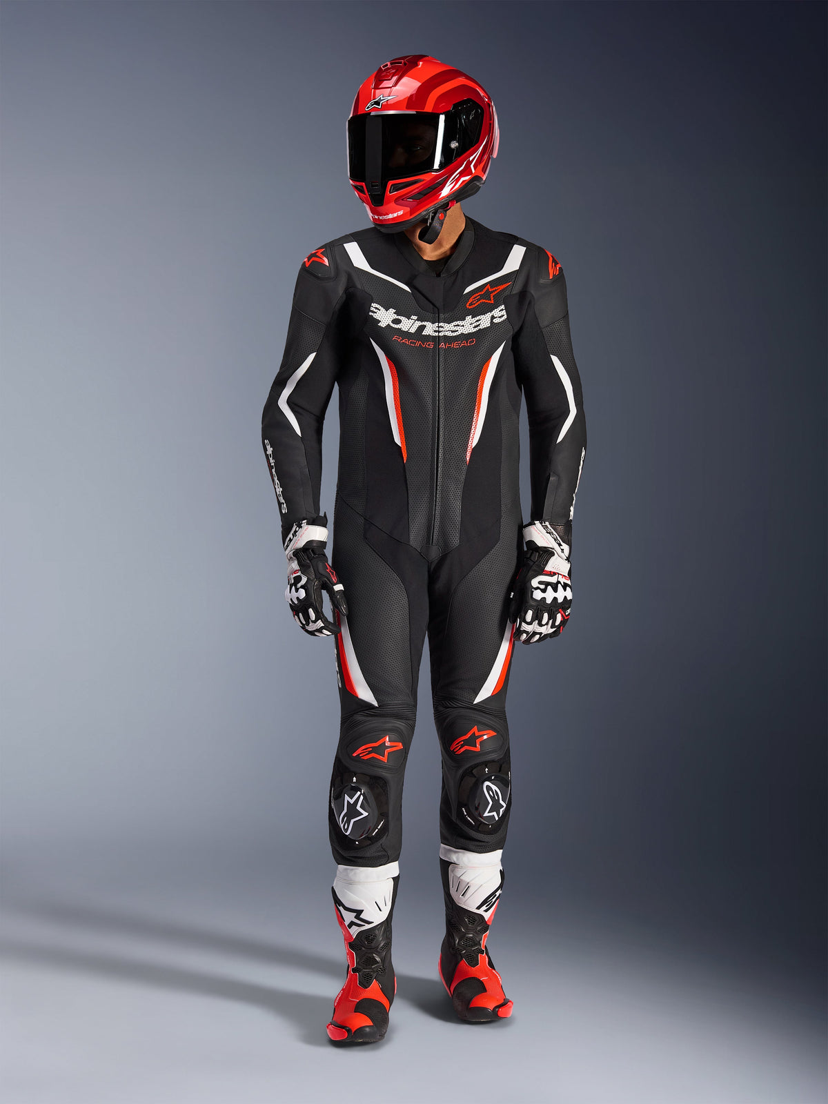 Tuta in pelle Alpinestars GP Force V2 - 1 pezzo, nero, bianco e rosso fluo, indossata con casco da corsa rosso, guanti bianchi e neri e stivali rossi e neri, con pannelli in pelle traforata e logo Alpinestars, abbigliamento professionale da gara motociclistica