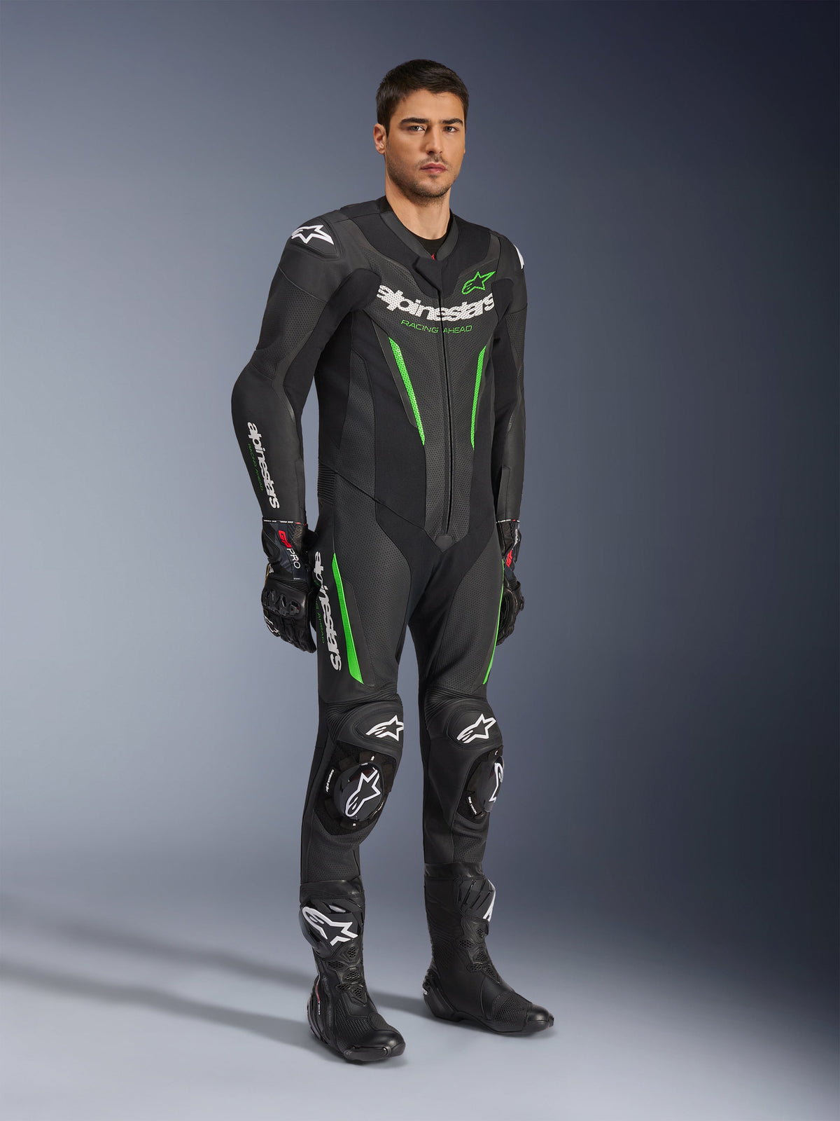 Tuta in pelle Alpinestars GP Force V2 intera, nera e verde, abbigliamento completo da corsa indossato con guanti e stivali neri, con pannelli in pelle traforata, marchio Alpinestars bianco e slider ginocchia integrati, tuta professionale per prestazioni in pista