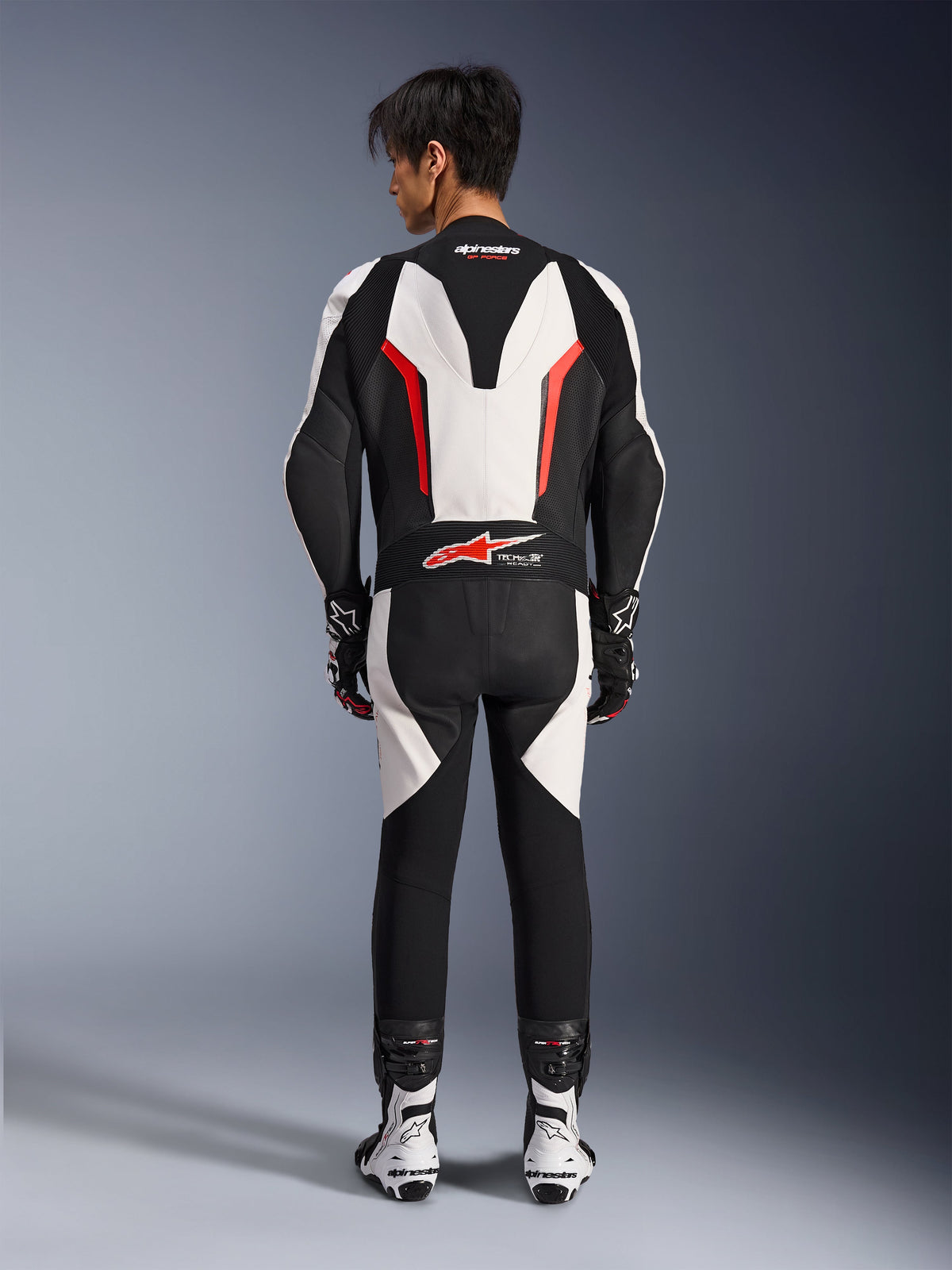 Alpinestars GP Force V2 Leather Suit 1-Piece, tuta da moto in pelle, bianco, nero e rosso fluo, vista posteriore del modello che indossa la tuta con stivali e guanti racing, con gobba aerodinamica, pannelli elastici a fisarmonica e marchio Tech-Air Ready