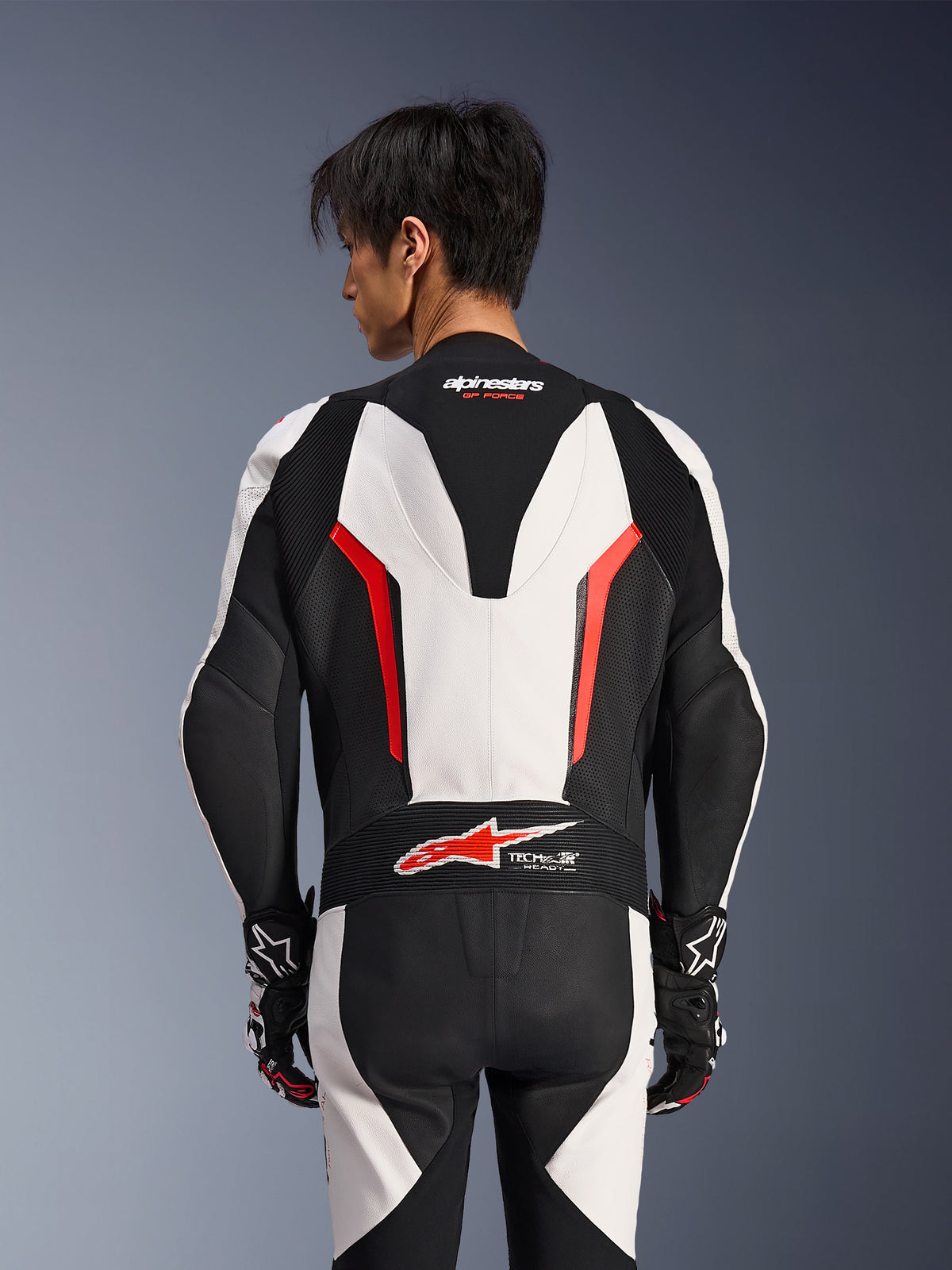 Alpinestars GP Force V2 Leather Suit 1 Pezzo, bianco, nero e rosso fluo, vista posteriore su modello maschile, con gobba aerodinamica, pannelli in pelle traforata e logo Tech-Air Ready sulla parte bassa della schiena, abbigliamento racing professionale con guanti neri e bianchi