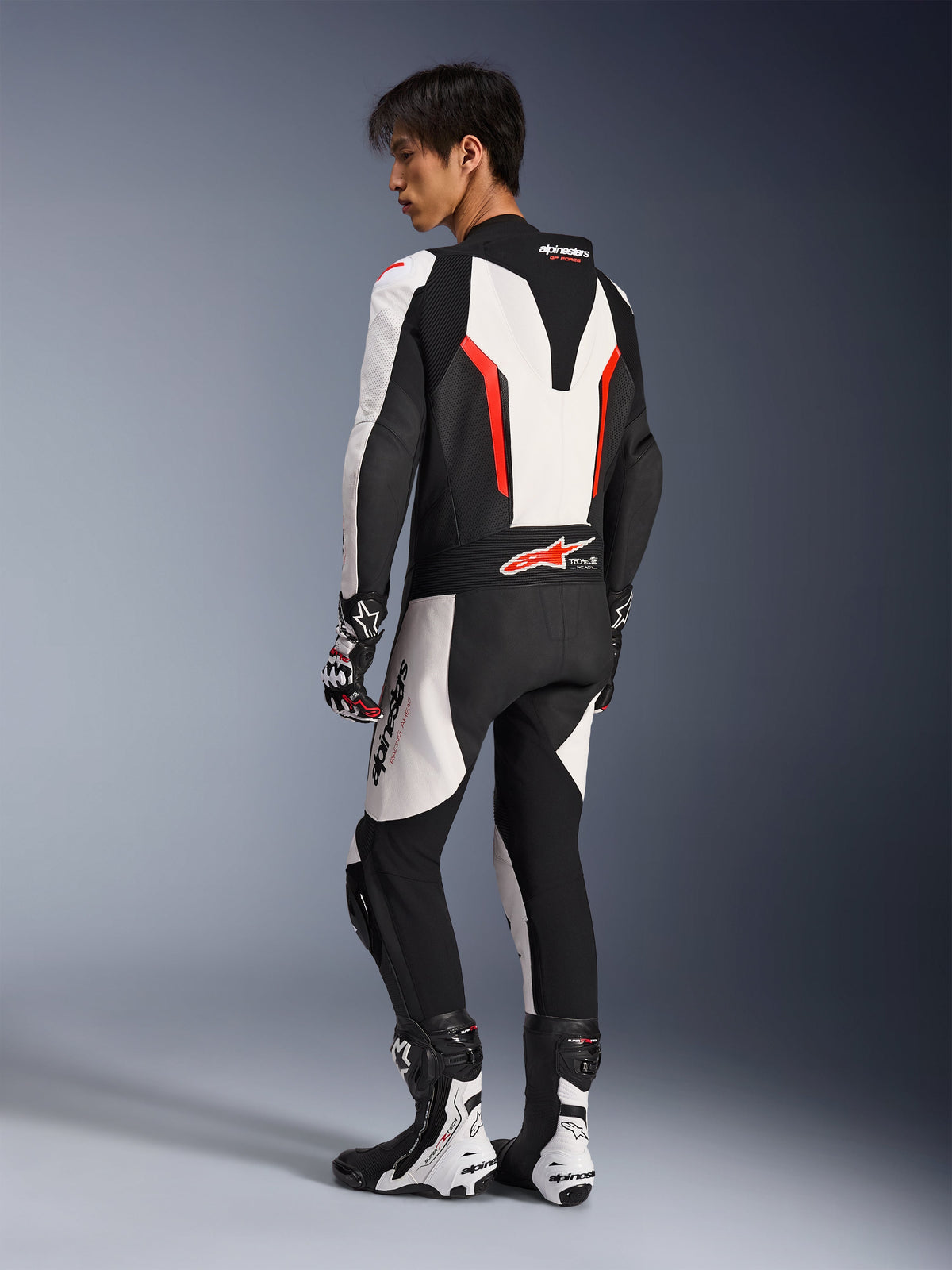 Pilota di moto che indossa la tuta in pelle Alpinestars GP Force V2 - 1 Pezzo in bianco, nero e rosso fluo, vista posteriore-laterale che mostra la gobba aerodinamica e i pannelli in pelle traforata, abbinata a stivali e guanti da corsa coordinati, progettata per la pista e la guida ad alte prestazioni.