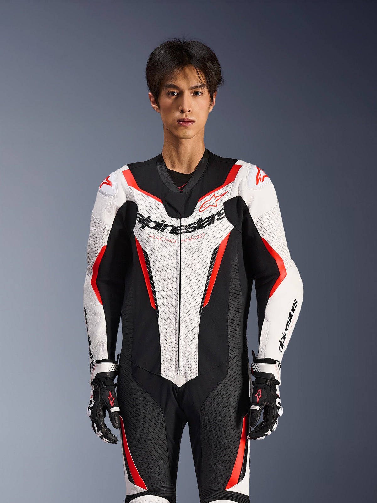 Tuta in pelle Alpinestars GP Force V2 - Monopezzo, bianco, nero e rosso fluo, indossata da un modello, con pannelli in pelle traforata e logo Alpinestars sul petto, coordinata con guanti da corsa bianchi e neri, progettata per la pista e la guida sportiva