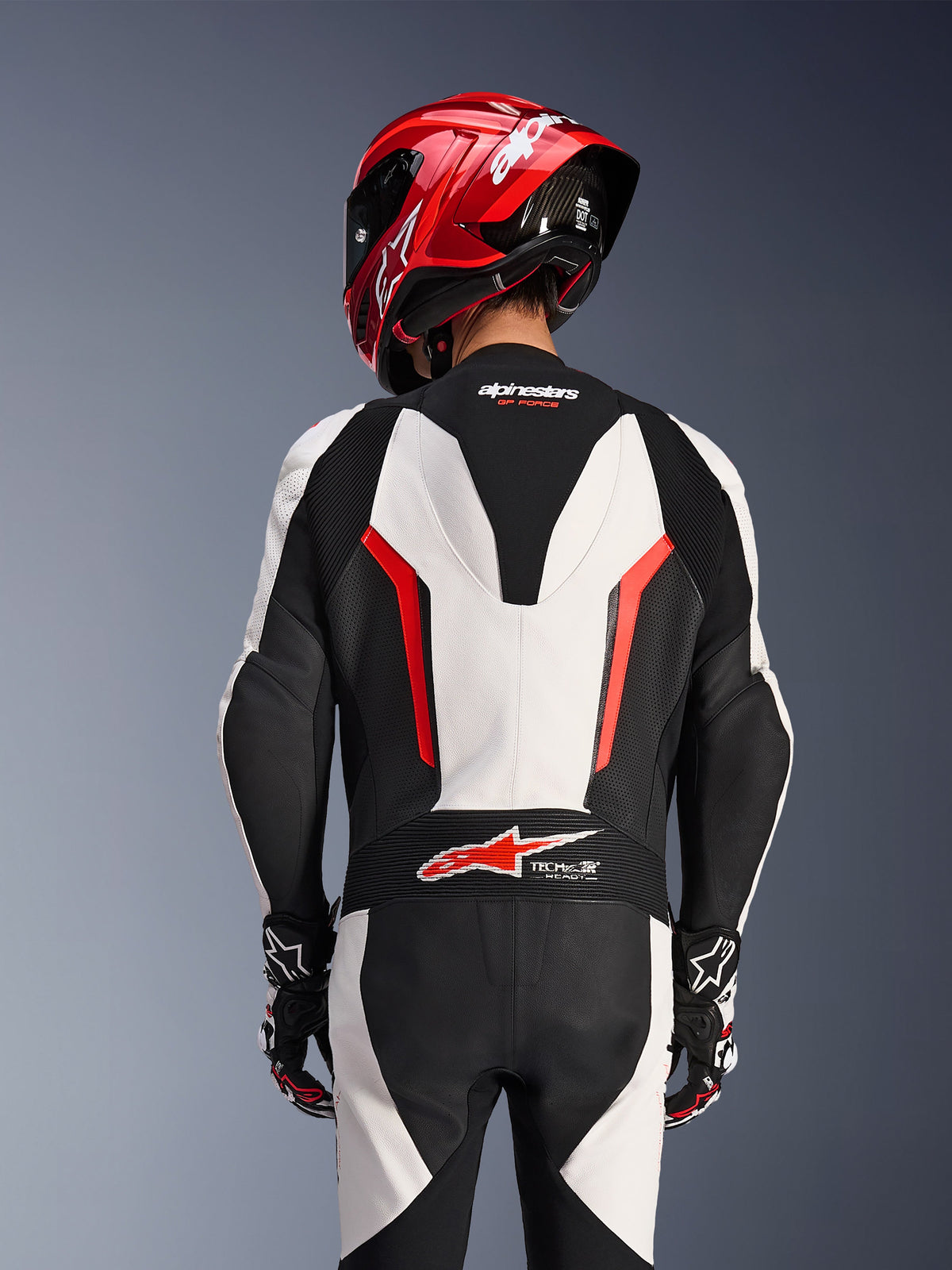 Vista posteriore della tuta in pelle monopezzo Alpinestars GP Force V2 in bianco, nero e rosso fluo, indossata con un casco da corsa rosso, con logo Tech-Air Ready sulla parte bassa della schiena, pannelli in pelle traforata e zone elasticizzate a fisarmonica per prestazioni racing.