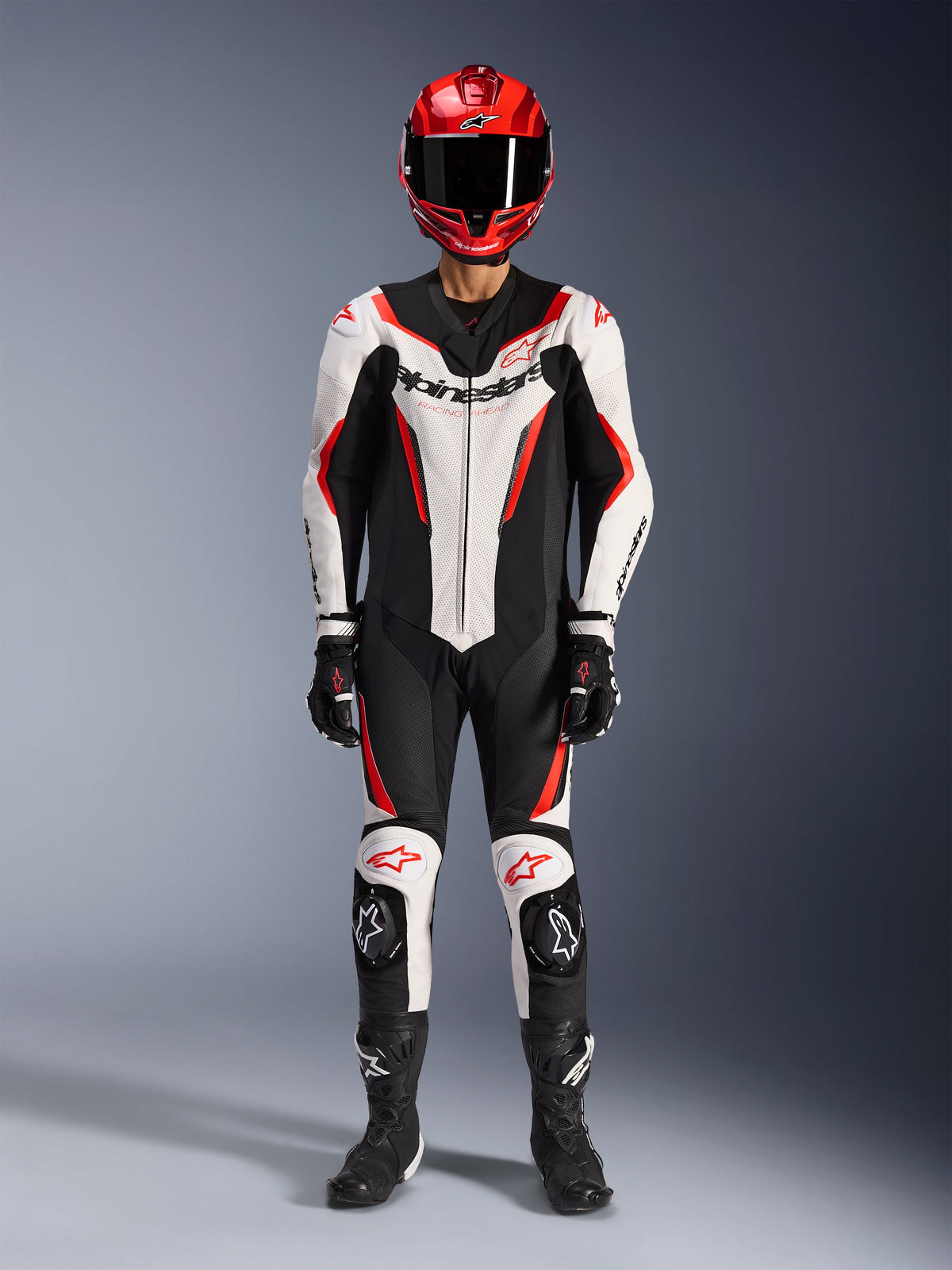 Tuta in pelle Alpinestars GP Force V2 monopezzo, bianco, nero e rosso fluo, equipaggiamento racing completo con casco rosso, guanti e stivali neri, caratterizzata da design aerodinamico e loghi Alpinestars su petto e ginocchia