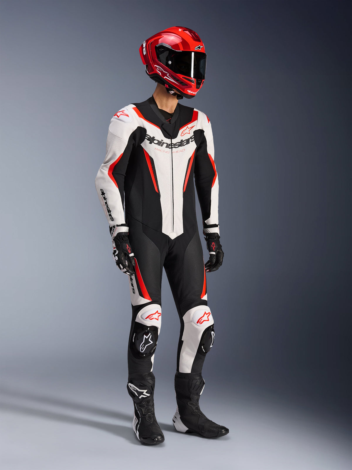 Alpinestars GP Force V2 Leather Suit 1-Piece in bianco, nero e rosso fluo, scatto in studio a figura intera di un pilota con equipaggiamento completo da gara, comprensivo di casco rosso, guanti neri e stivali neri, costruzione aerodinamica in pelle bovina con loghi e slider per ginocchia integrati.