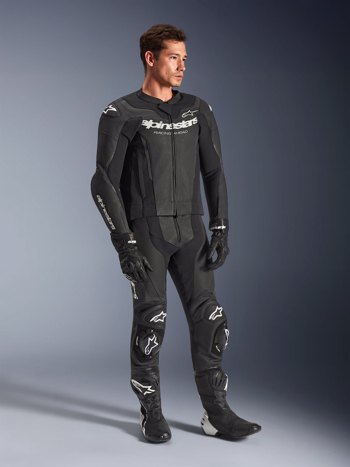 Tuta in pelle Alpinestars GP Force V2 2 pezzi, nero, indossata da un modello con guanti e stivali da corsa neri coordinati, con marchio Alpinestars bianco, pannelli in pelle traforata per la ventilazione e saponette integrate, abbigliamento professionale da gara motociclistica