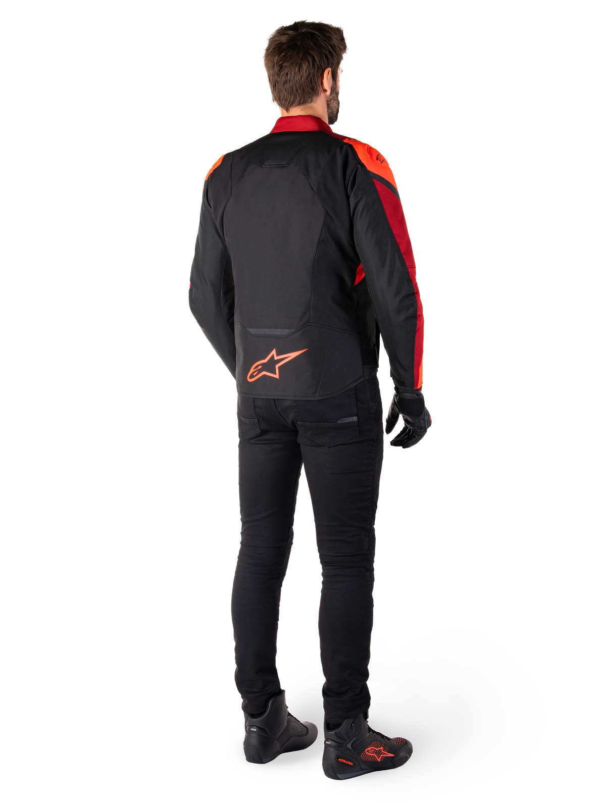 Alpinestars T-Jaws V4 Waterproof Jacket in Nero Rosso Fluo Rosso Scuro, vista posteriore di un motociclista che indossa la giacca in tessuto con pantaloni neri e stivali da guida, con logo Alpinestars rosso sulla parte bassa della schiena.