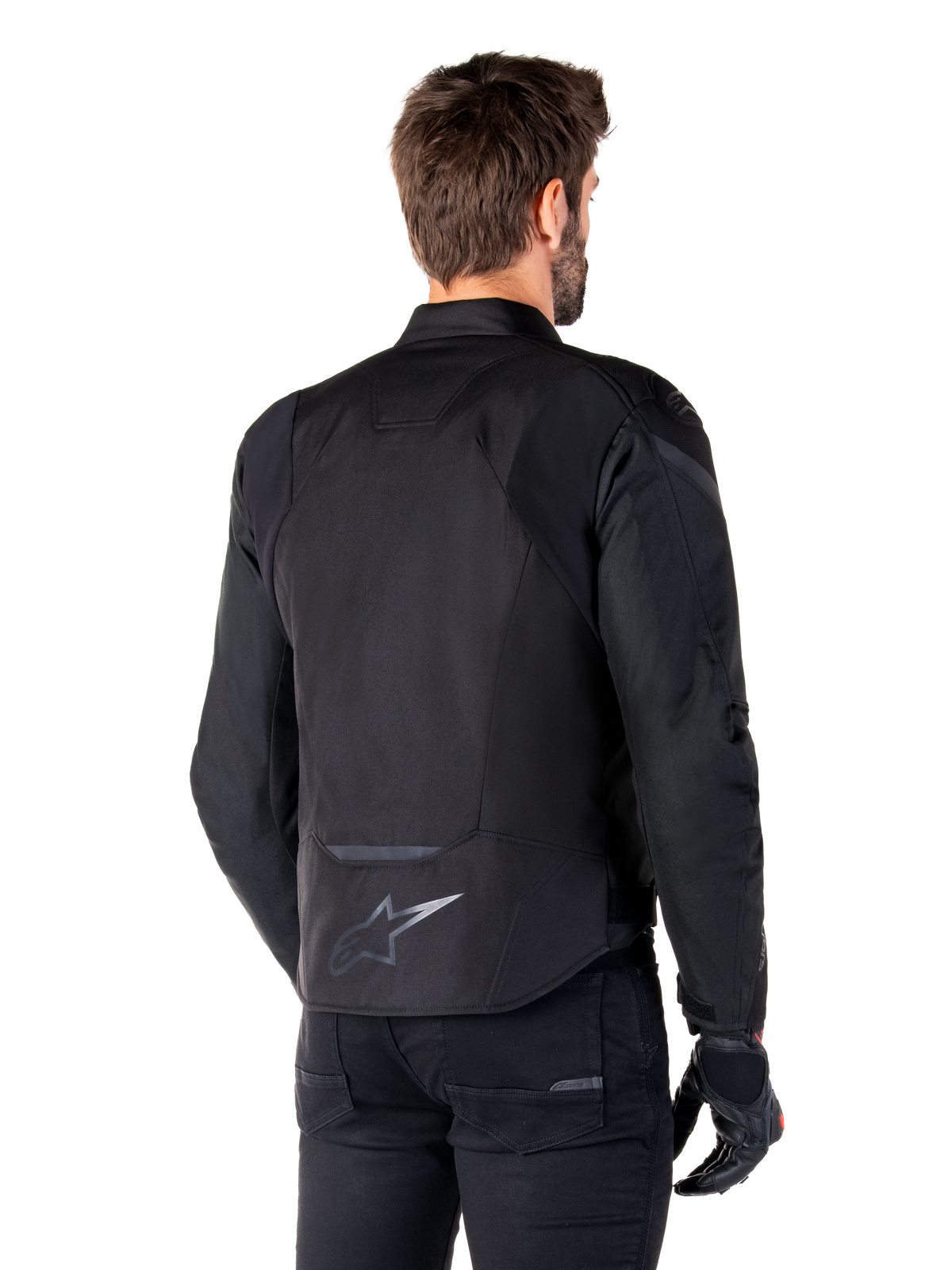 Giacca Alpinestars T-Jaws V4 Waterproof, nero, vista posteriore su modello, con logo Alpinestars riflettente sulla parte bassa della schiena, dorso allungato per una maggiore copertura, design sportivo in tessuto con pannelli tono su tono, indossata con guanti da moto neri