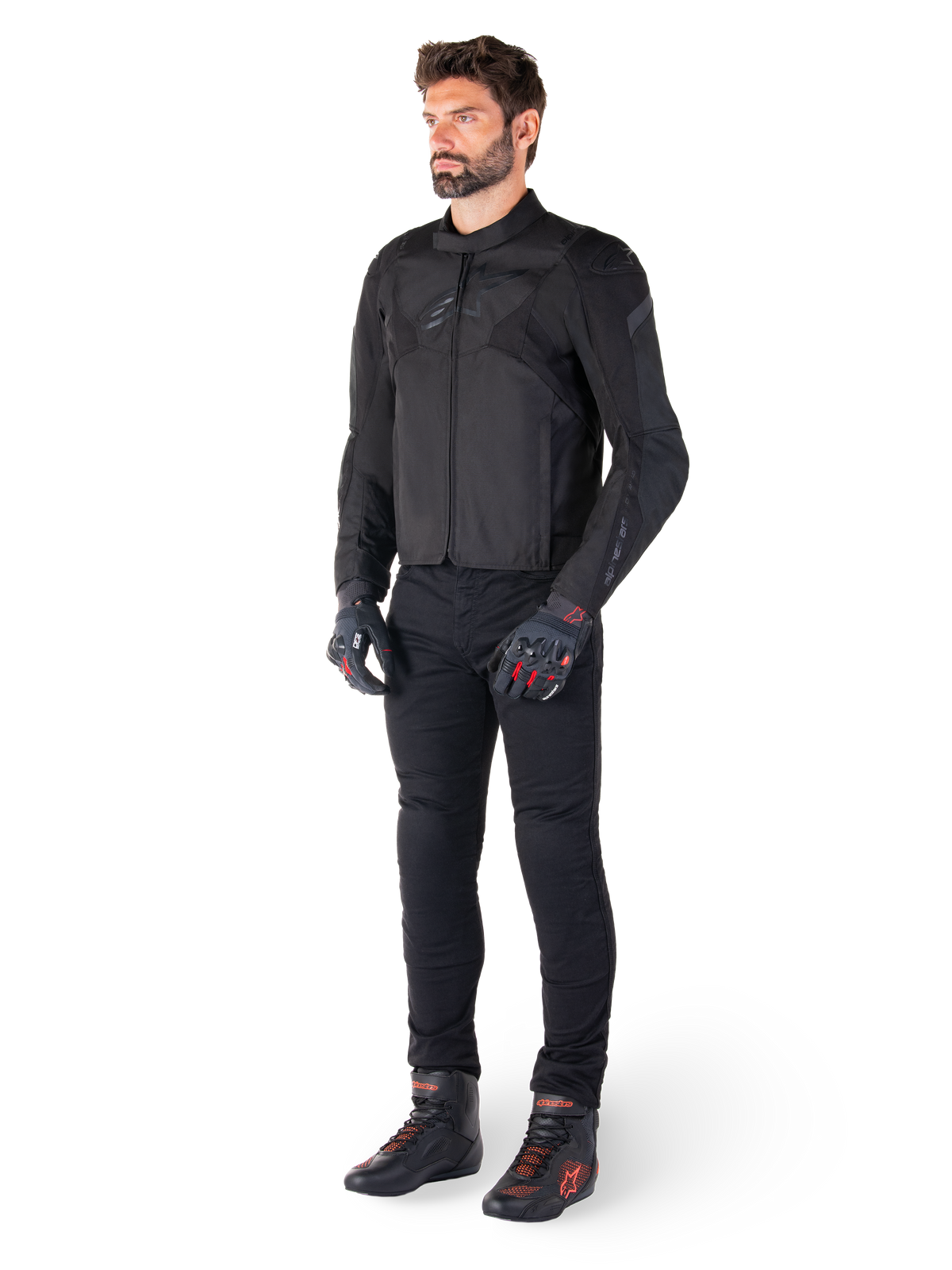 Modello maschile che indossa la giacca Alpinestars T-Jaws V4 Waterproof in nero, abbinata a pantaloni da guida neri, guanti neri con accenti rossi e scarpe da moto nere, con una vestibilità sportiva e loghi discreti, isolato su sfondo bianco.